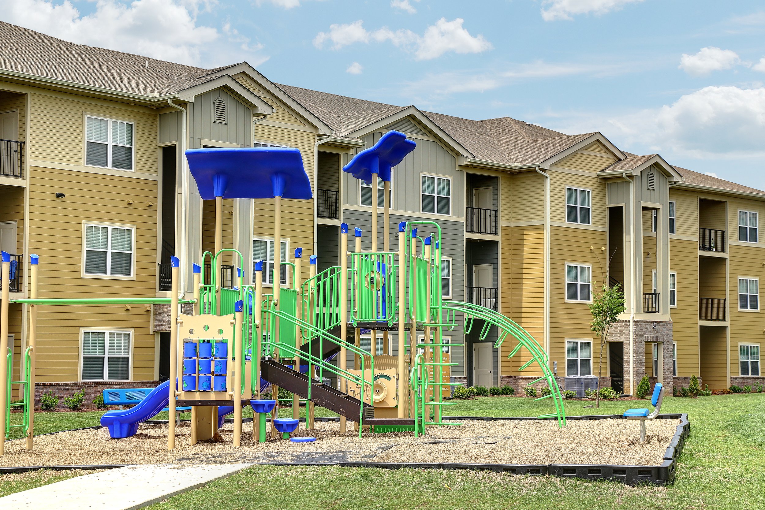 Colorful playground with slides, climbing structures, and swings in front of a yellow residential building with multiple floors and balconies.