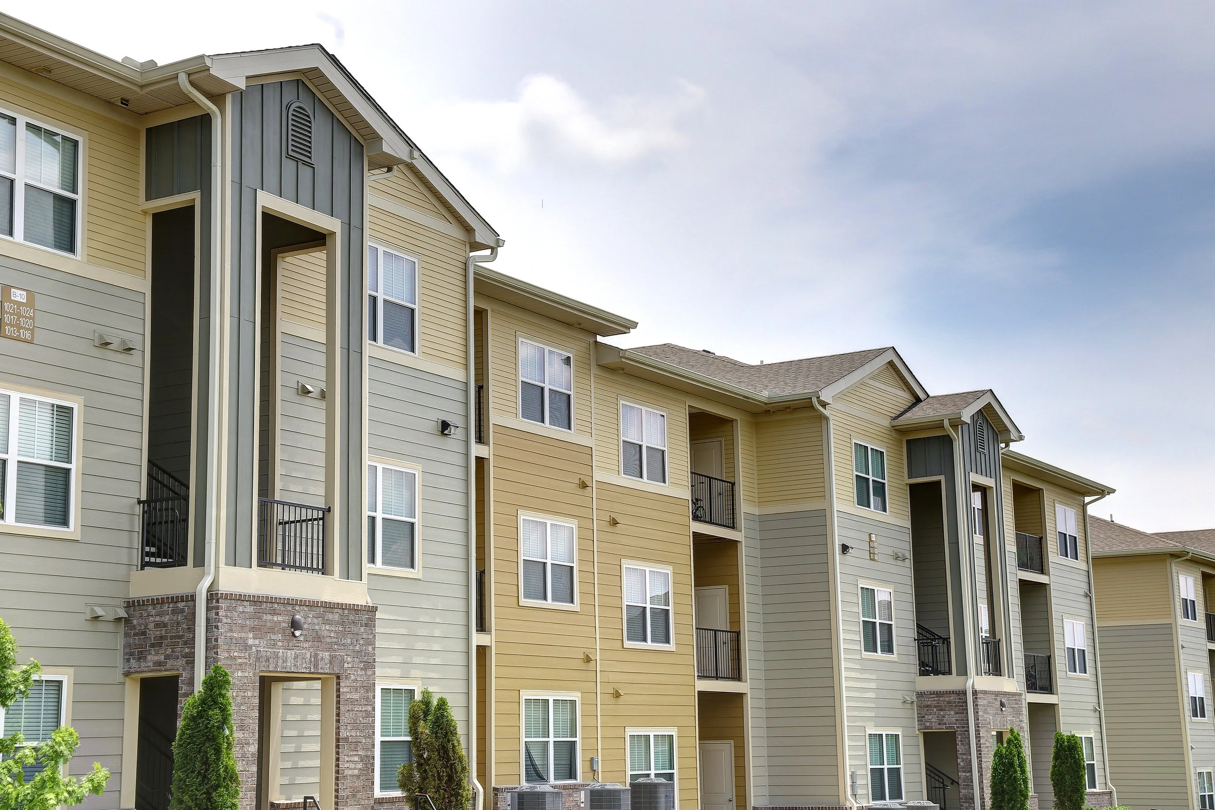 Multiple stories of a residential apartment complex with yellow and beige siding, brick accents at the base, and several windows. Small trees and bushes are in front.