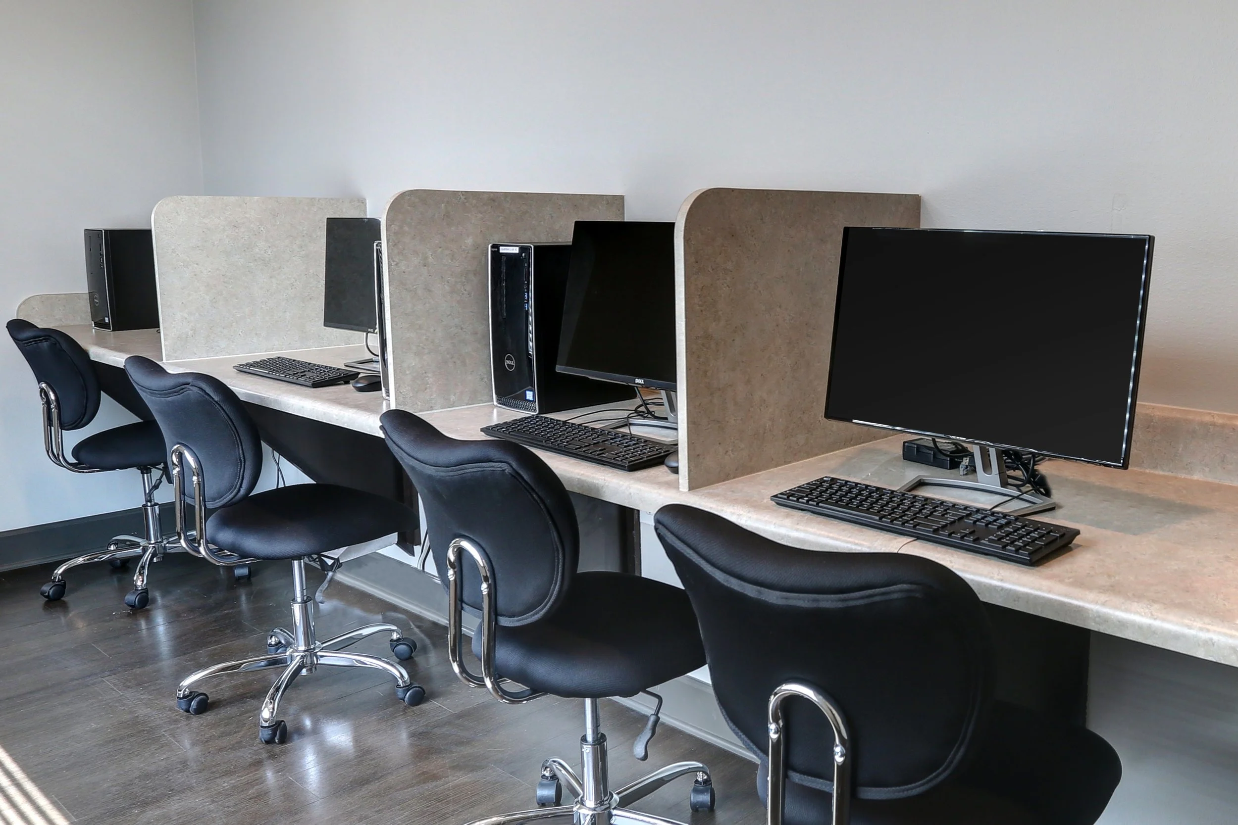 A row of computer desks with black chairs in an office, each station separated by beige divider panels, two desktop computers and monitors, keyboards, and mice on the desks.