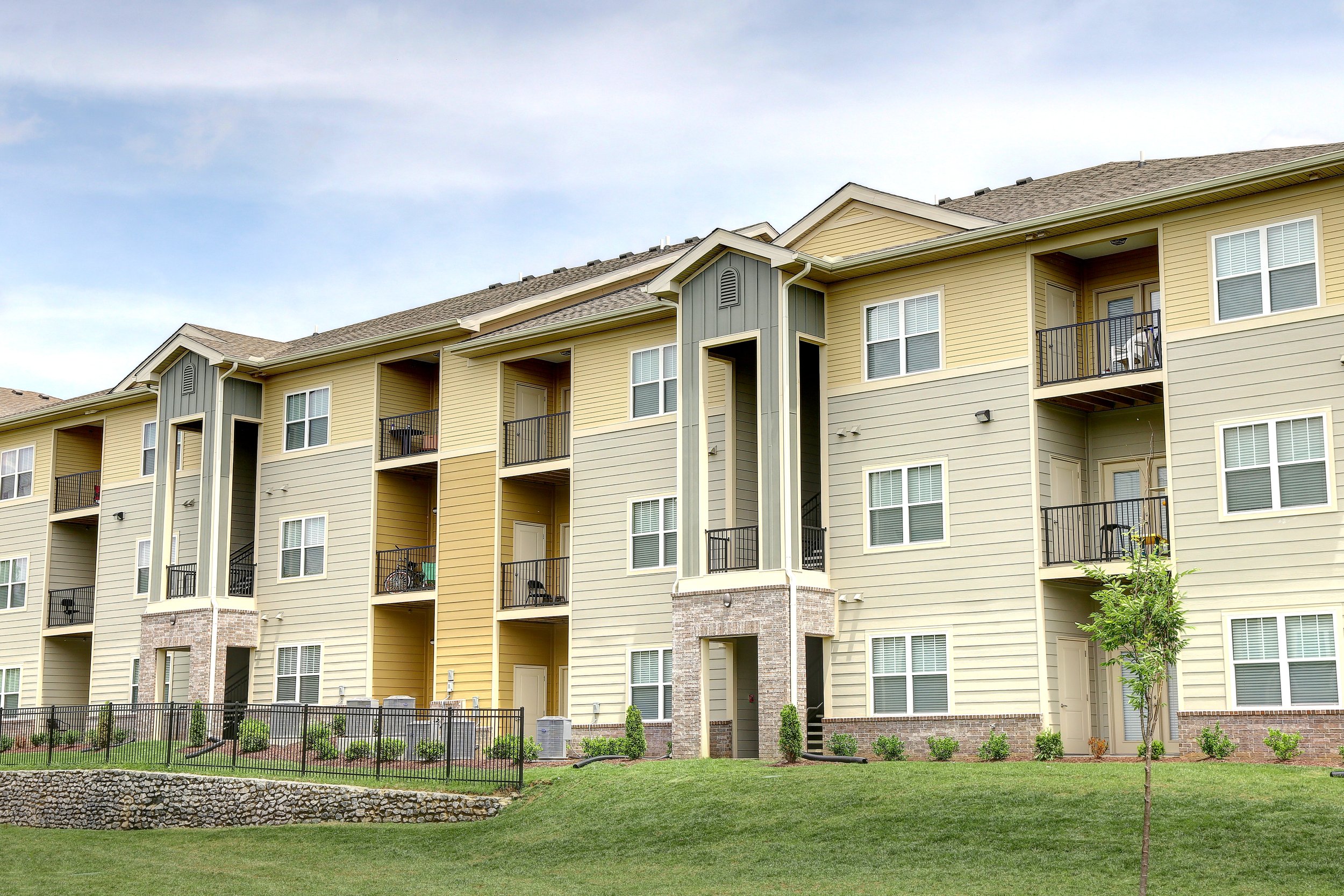 Multi-story residential building with balconies, beige and green exterior, and a small landscaped yard with a young tree in the foreground.