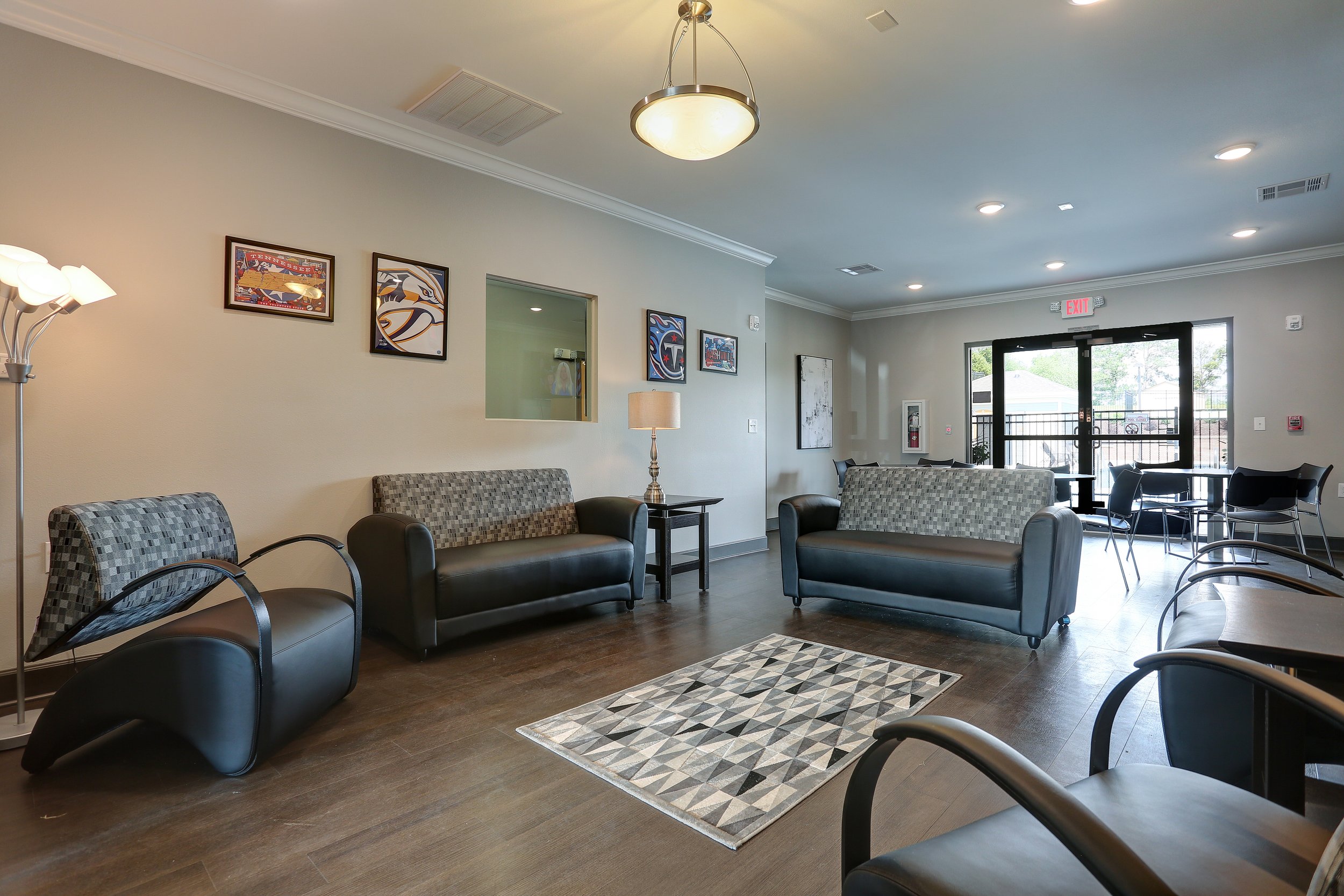 Lobby area with sofas, chairs, framed sports art on the wall, and a patterned rug on dark wood floor. Large window and glass door at the back.