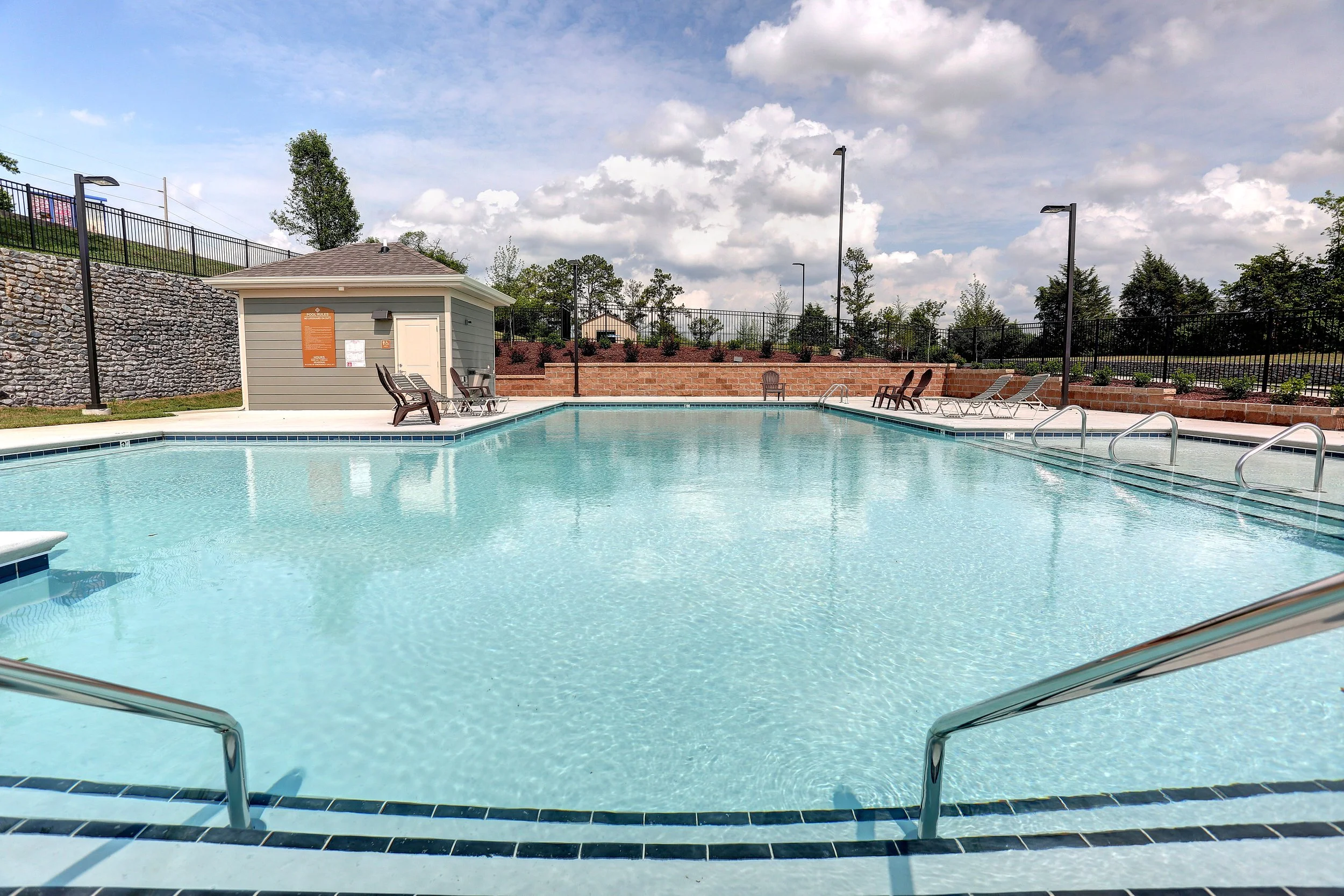 An outdoor swimming pool surrounded by lounge chairs, a small building, and a brick wall with trees and a cloudy sky in the background.