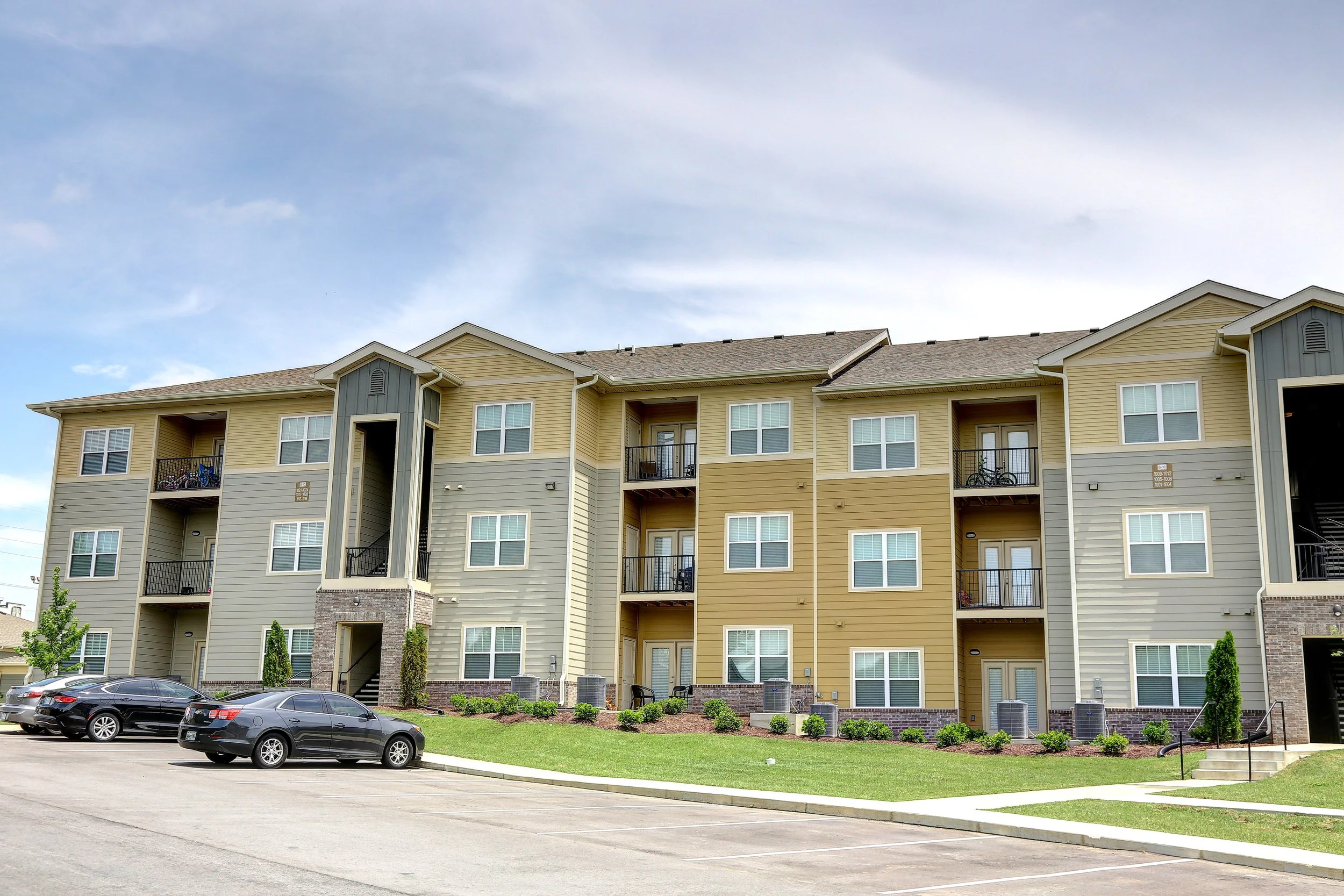 Exterior view of a multi-story apartment complex with yellow and gray siding, small balconies, and a parking lot in front with several cars, under a partly cloudy sky.