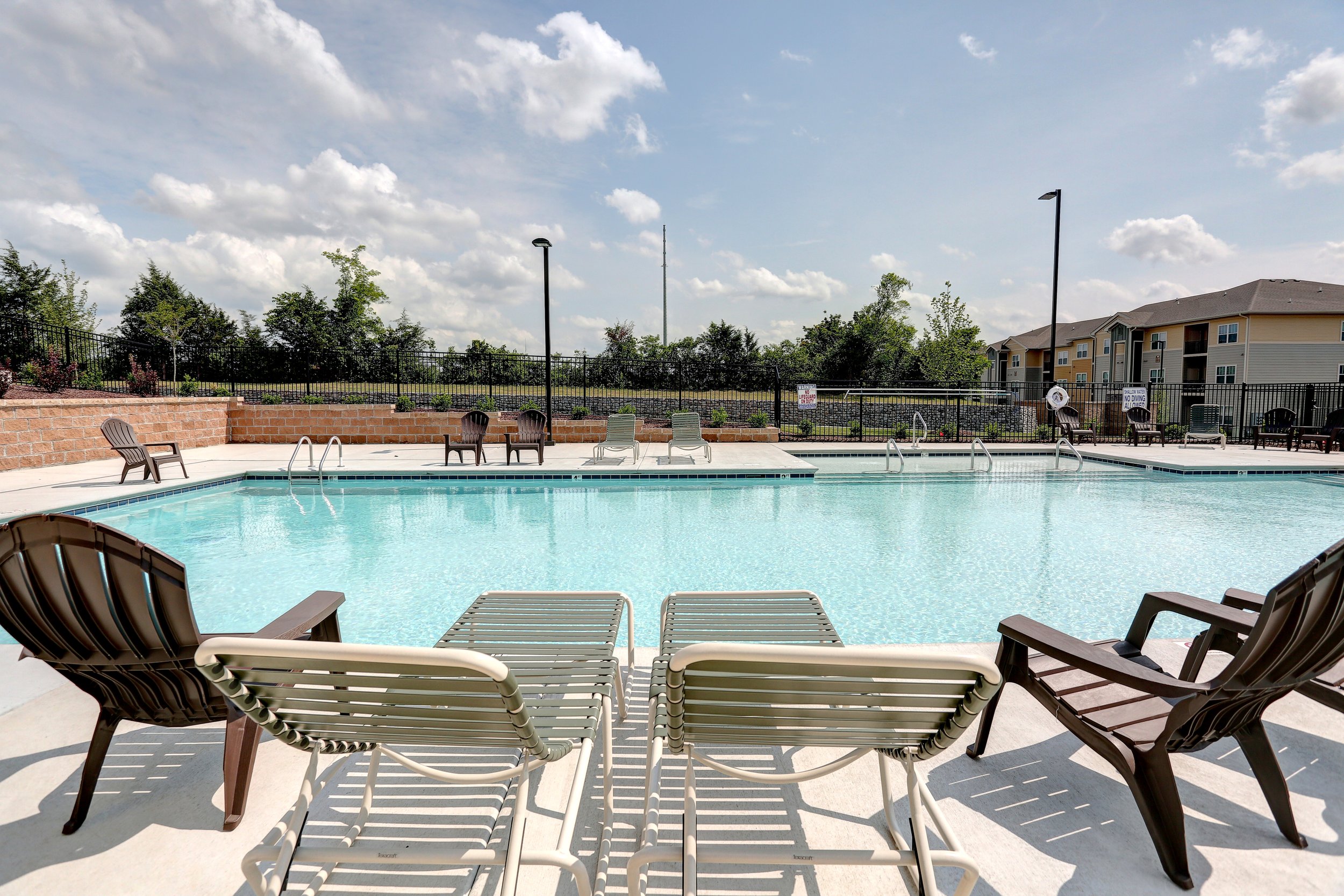 An outdoor swimming pool surrounded by lounge chairs and a black metal fence, with a row of residential buildings and trees in the background under a partly cloudy sky.