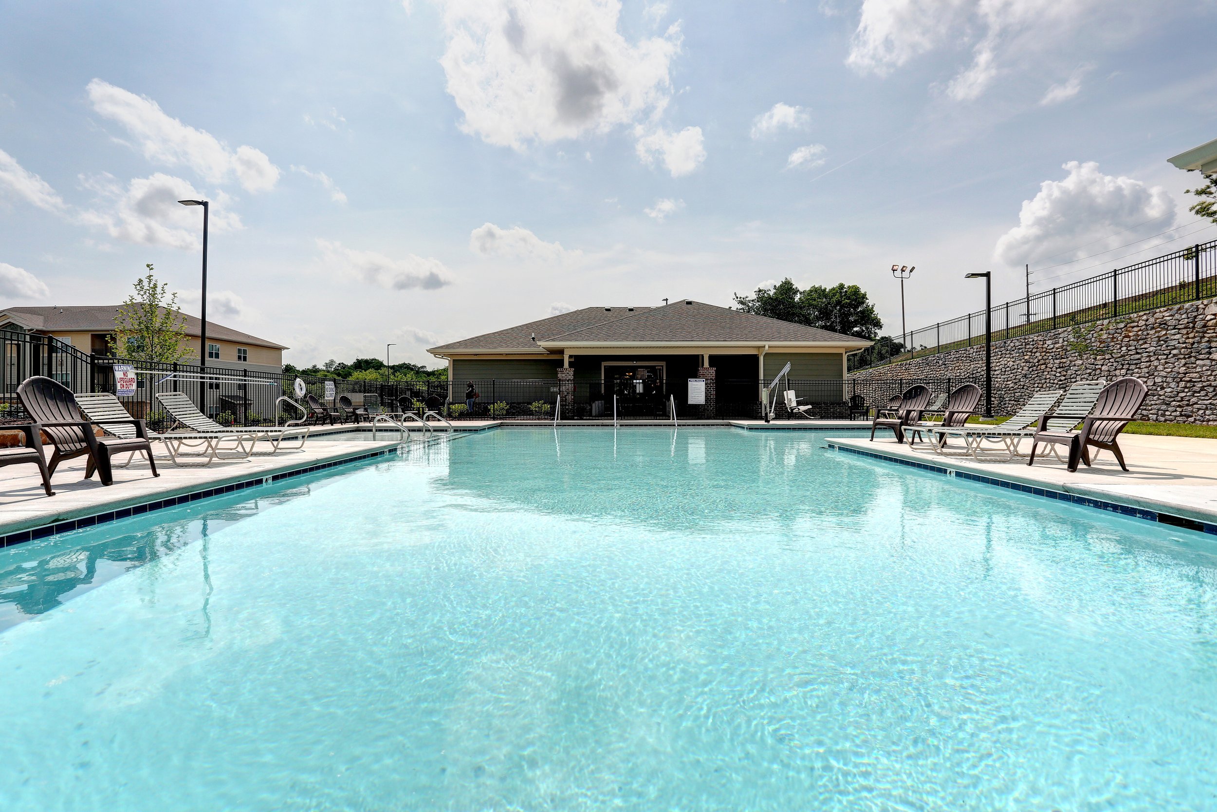 An outdoor swimming pool surrounded by lounge chairs, with a building and a stone wall fence in the background. The sky is partly cloudy.