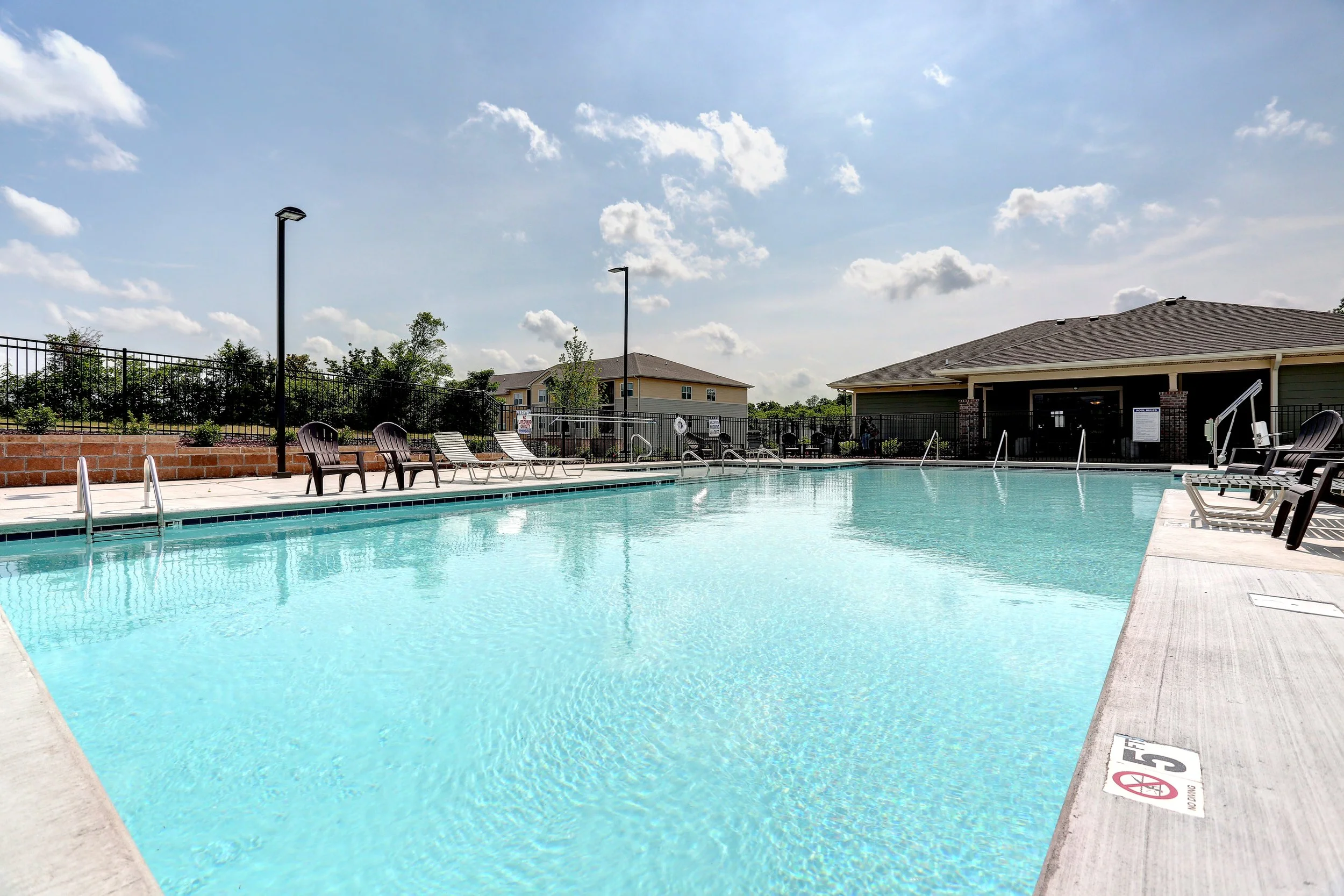 An outdoor swimming pool with clear blue water, surrounded by lounge chairs, a brick wall, and a fenced area with trees and residential buildings in the background. The sky is partly cloudy.