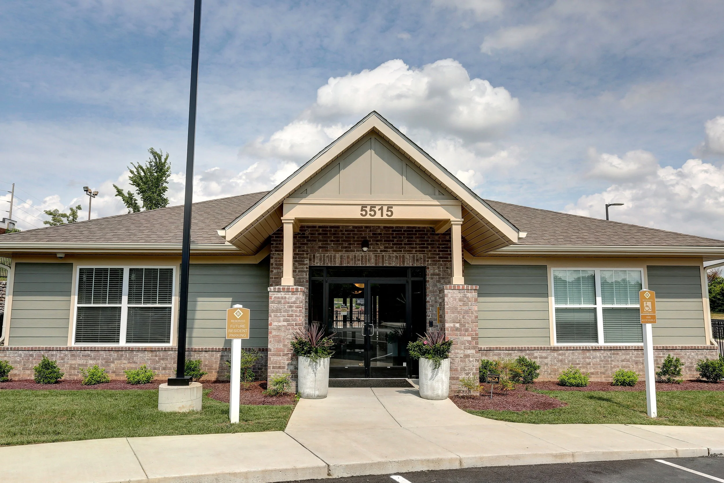 A newly constructed residential building with a gable roof and brick accents, featuring outside signs for reserved parking and handicapped accessibility, with a walkway leading to the entrance and landscaped bushes in front.