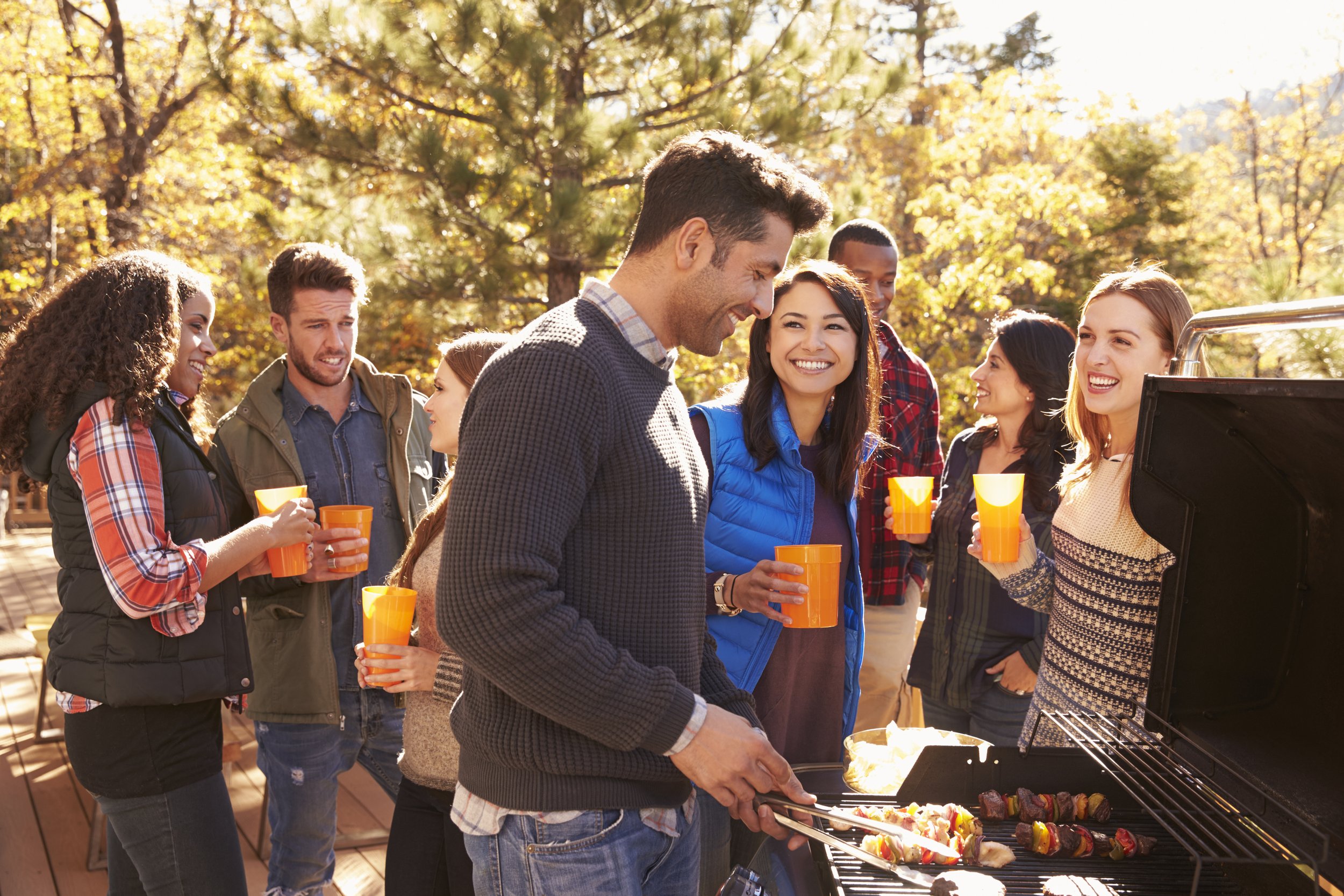 Group of people at an outdoor barbecue enjoying food and drinks in a park during autumn. Men and women are gathered around a grill with skewered meats and vegetables, some holding orange cups, smiling and socializing.