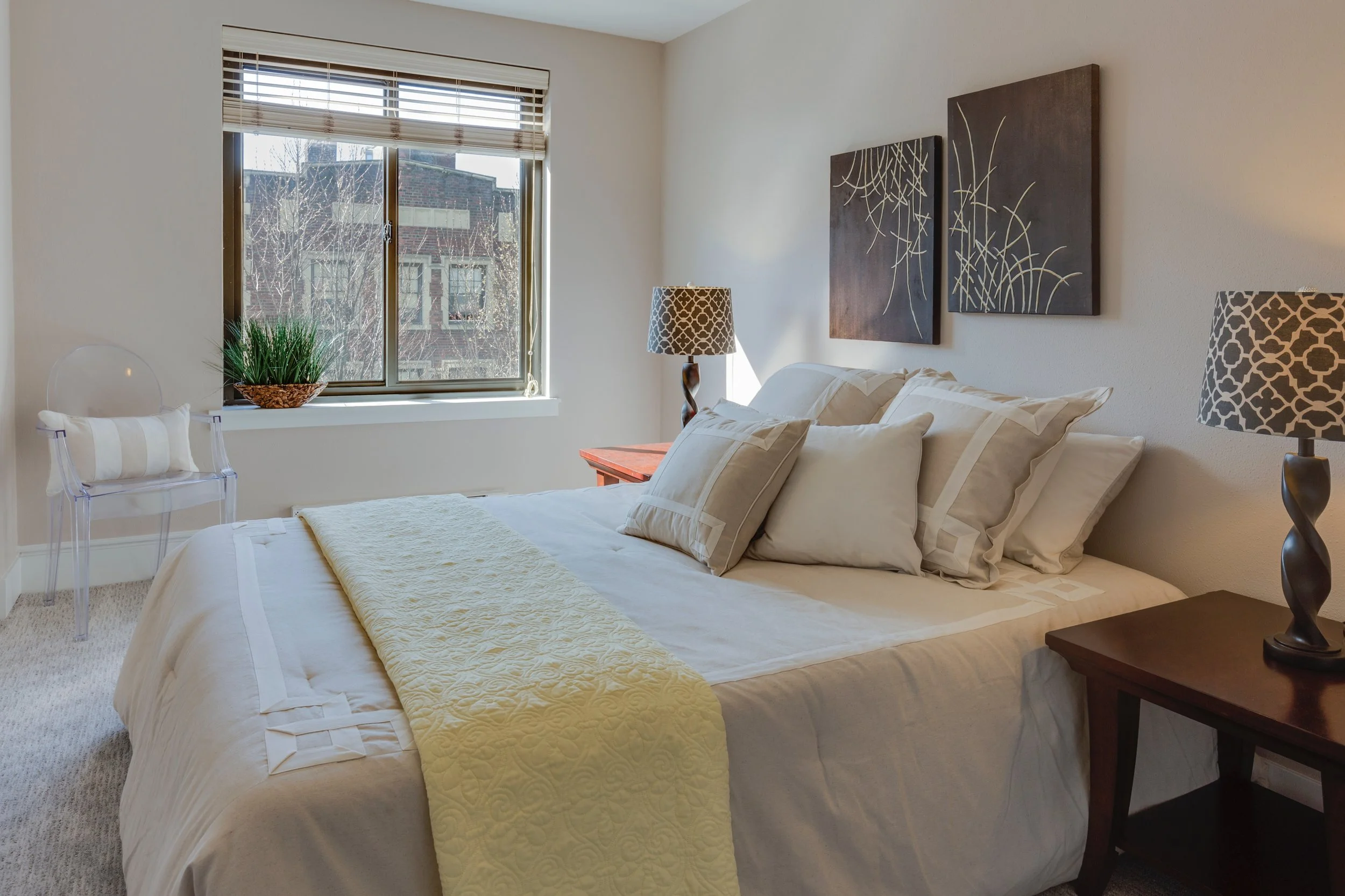A neatly made bed with cream-colored sheets and pillows, a yellow quilted throw, and two black and white patterned lamps on dark wood bedside tables in a bright bedroom with a window showing neighboring buildings.
