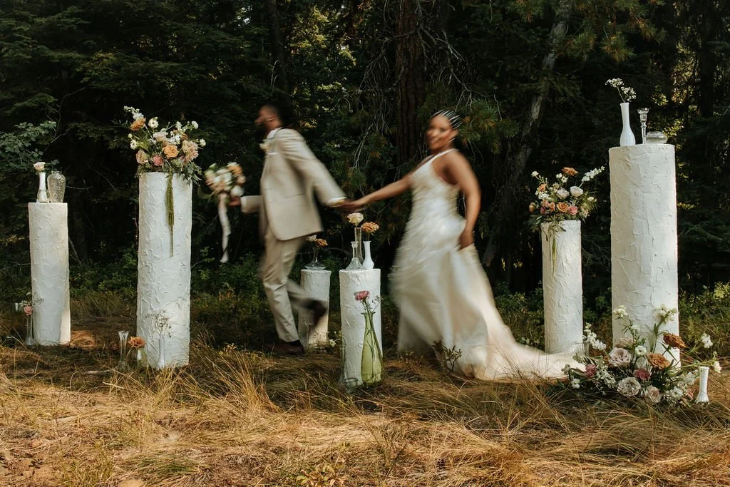 A couple in wedding attire walking hand-in-hand outdoors among white decorated pillars with flowers at a forest setting.
