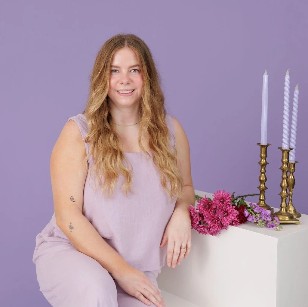 A woman with long hair, wear a light purple set, sitting next to a white pedestal with pink flowers and three tall purple candles in gold candleholders, against a purple background.