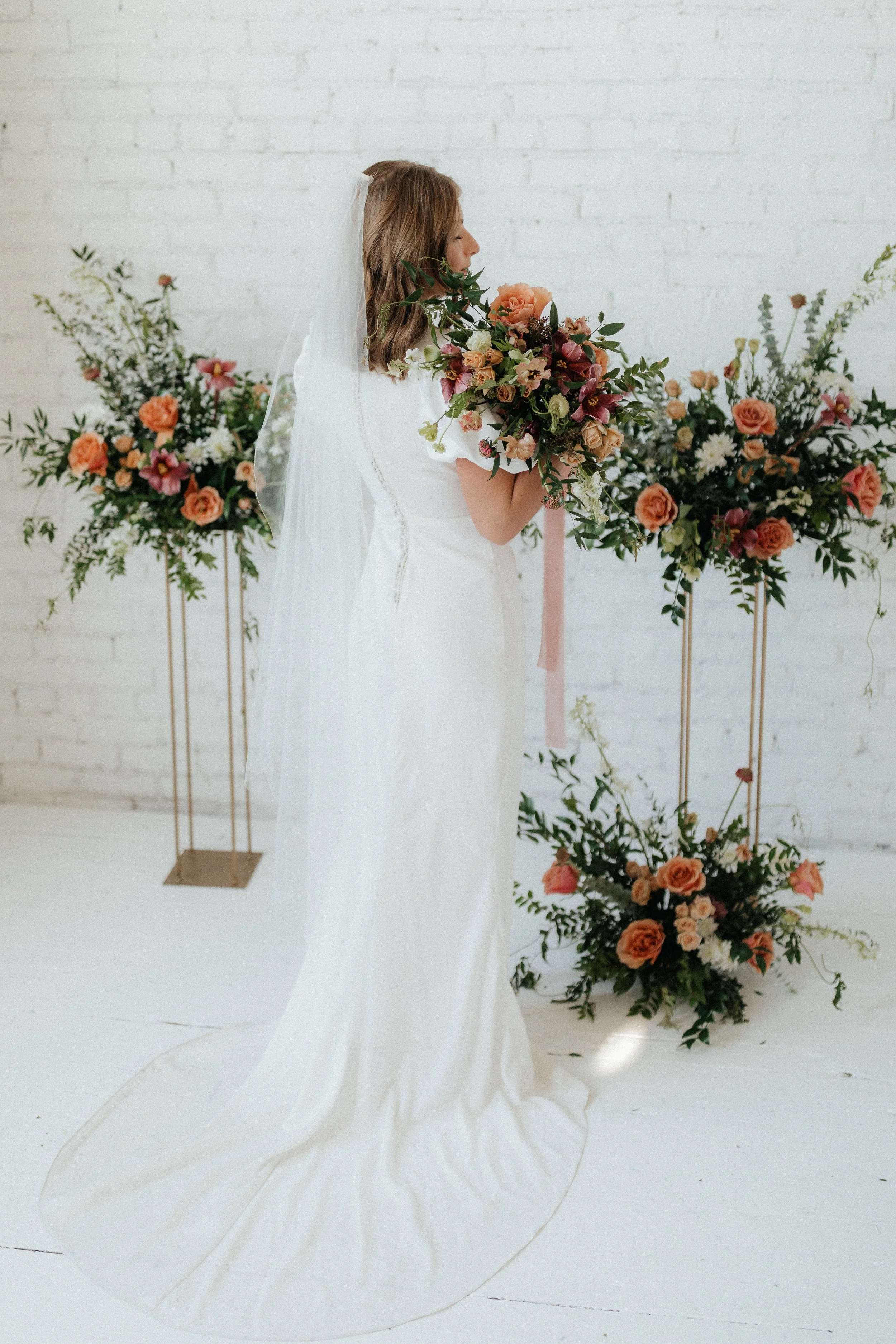 A bride holding a bouquet of flowers in front of her face, standing in front of floral arrangements against a white brick wall.
