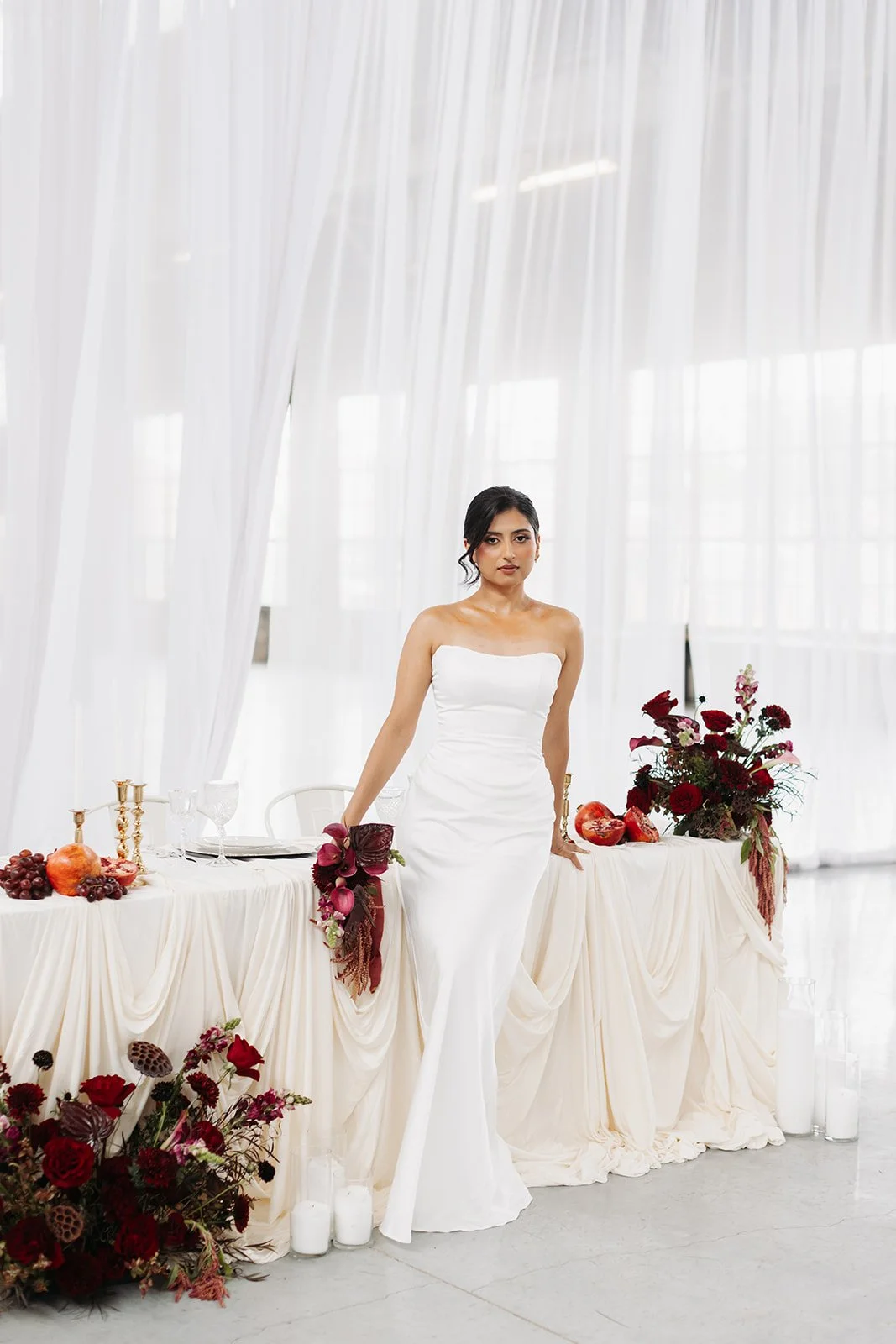 A woman in a white strapless wedding dress standing at a decorated table with red flowers and fruit in a bright, airy venue with white curtains.