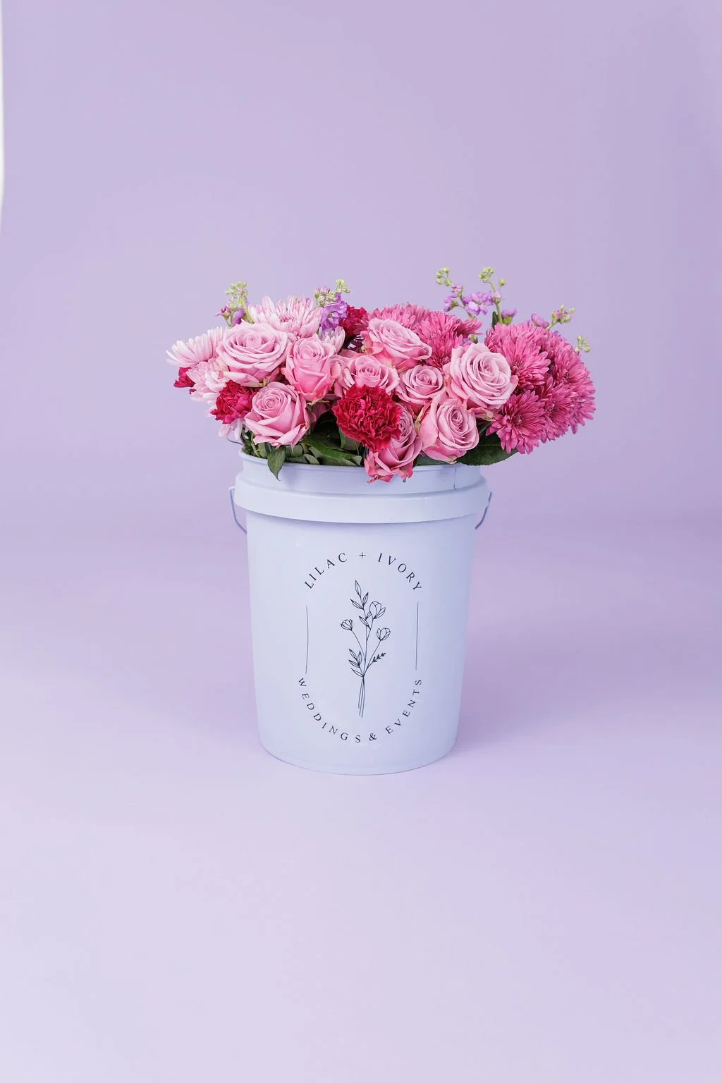 A white bucket with a logo reading 'Lilac + Ivory Weddings & Events' holds a bouquet of pink and purple flowers, including roses, carnations, and chrysanthemums, against a light purple background.