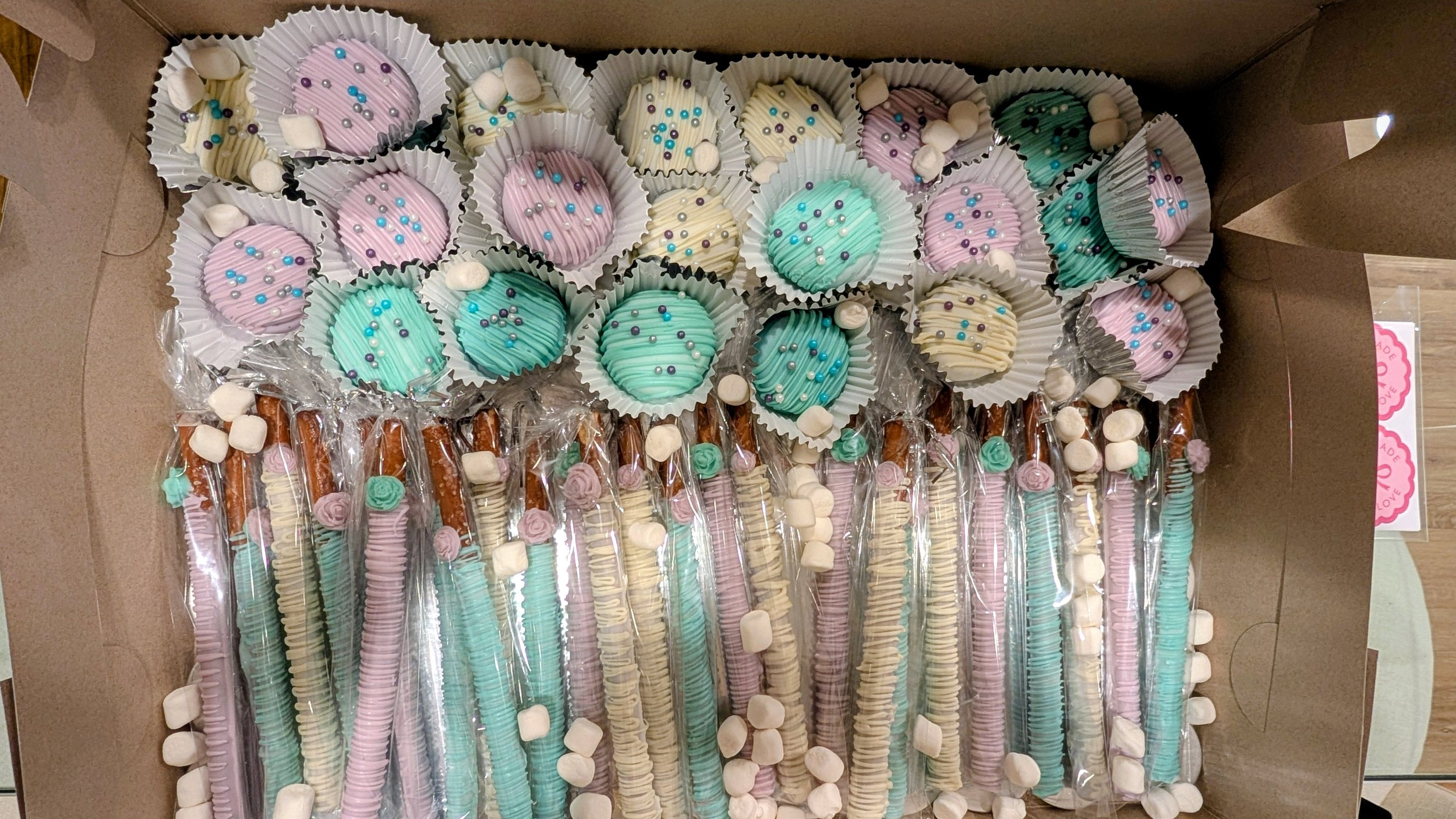 Display of pastel-colored candies and treats in a box, including marshmallow pops, chocolate-covered pretzels, and decorated marshmallow sweets, arranged in decorative cupcake liners.
