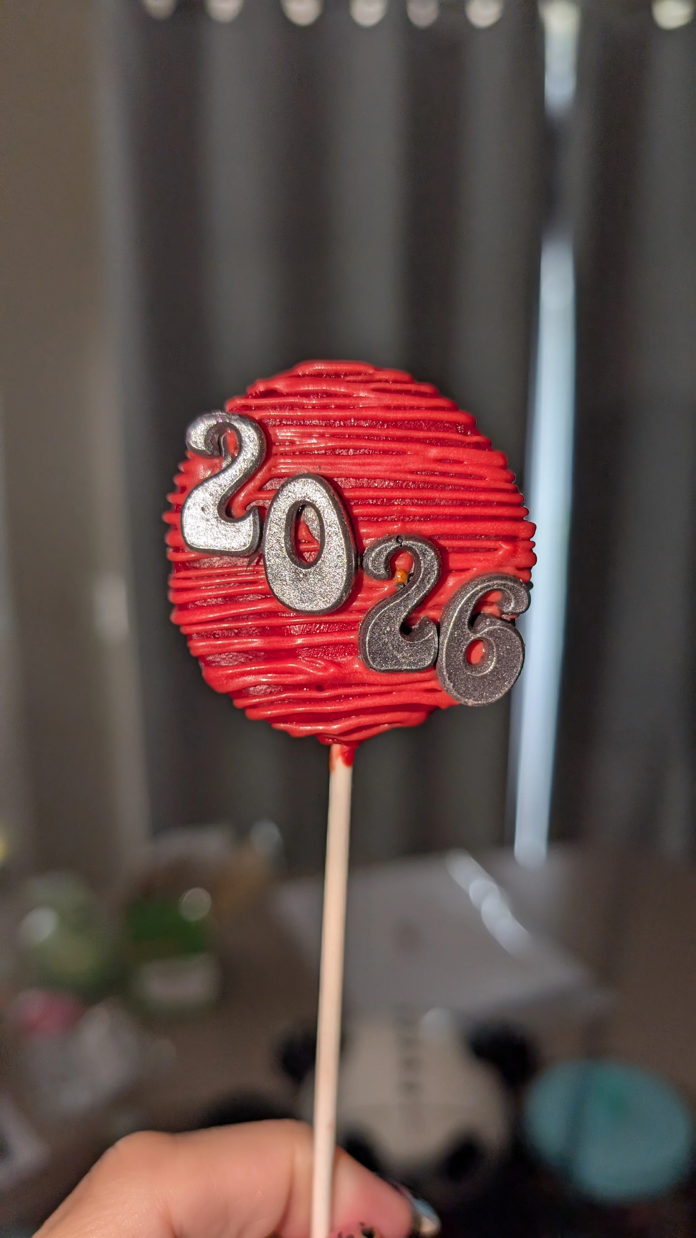 Red spherical cake pop decorated with the silver numbers '2026' on the front, on a white stick held by a person's hand.