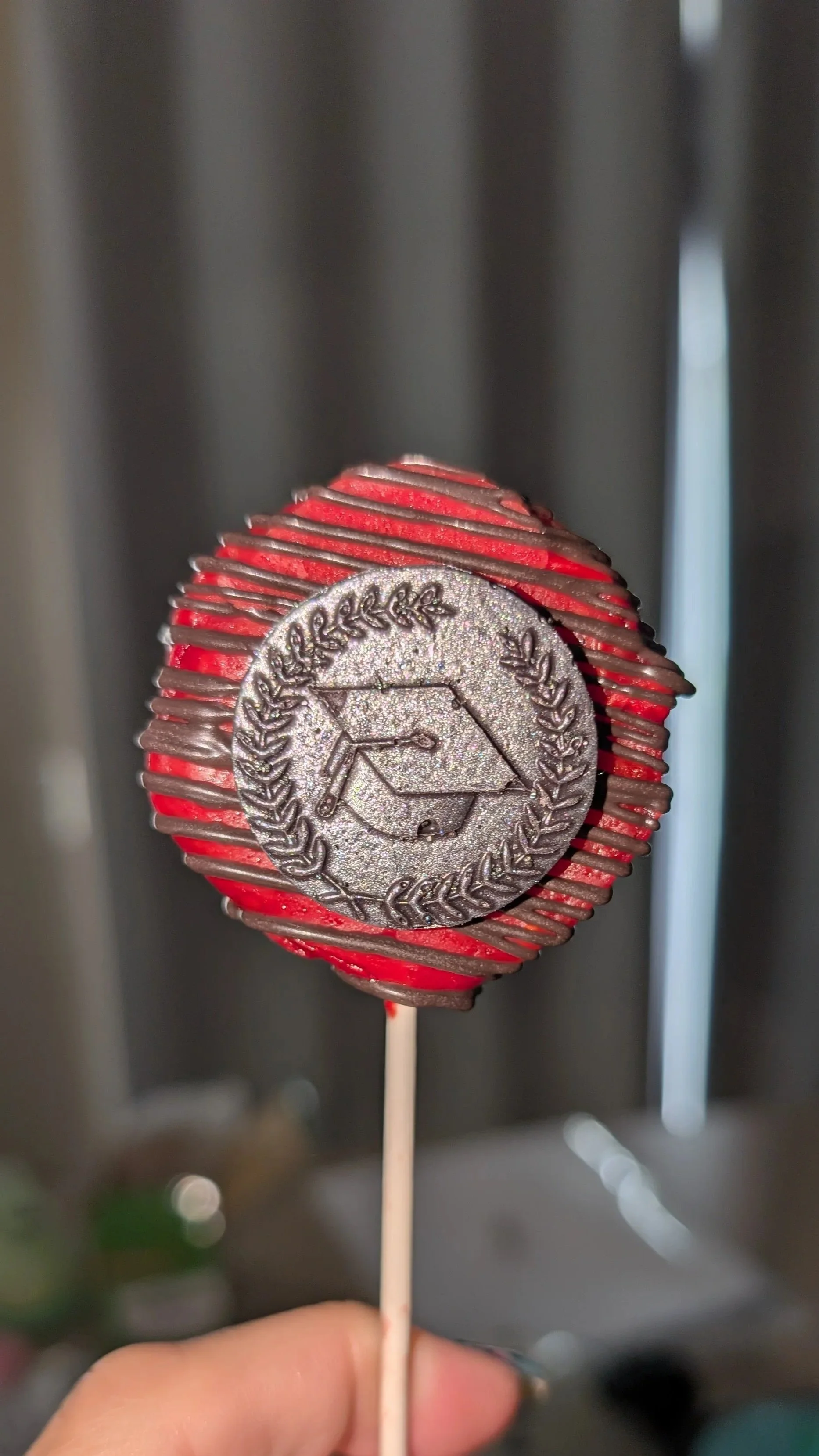A hand holding a cake pop with a silver medal and red and dark swirl pattern.