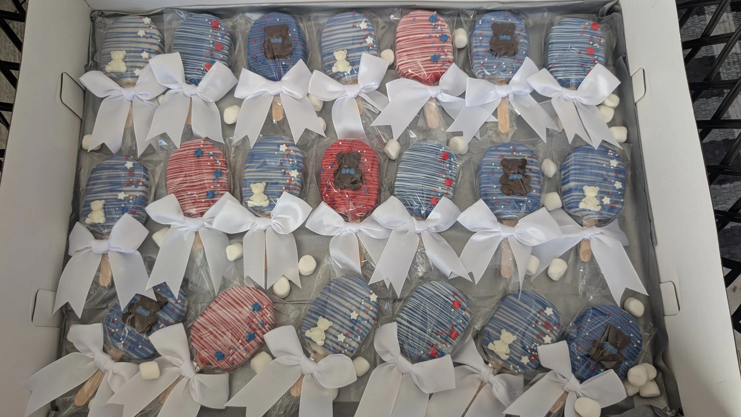 A box of decorated cookies with patriotic red, white, and blue themes, wrapped in clear plastic and tied with white ribbons, with small marshmallows scattered underneath.