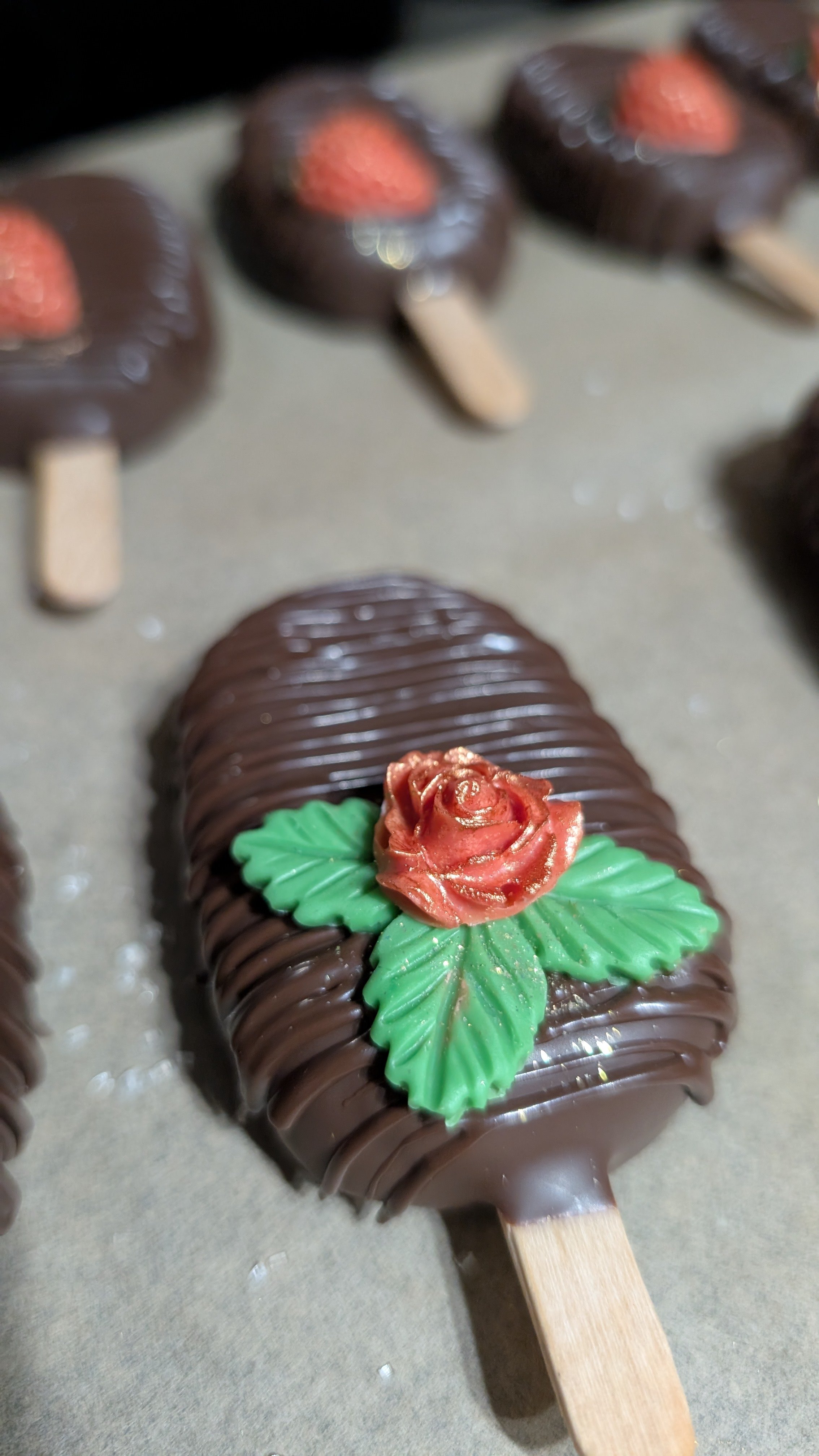 Chocolate-covered cakesicle with a red edible flower and green leaves decoration.