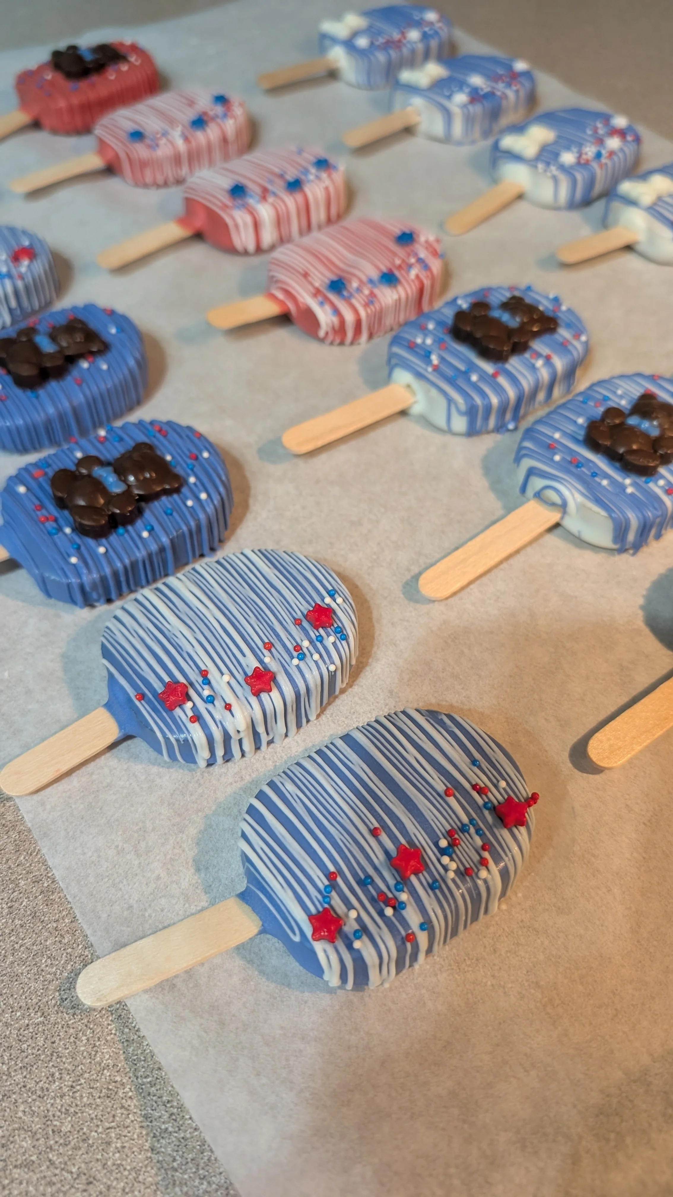 Patriotic-themed popsicles decorated with red, white, and blue icing, sprinkles, and star-shaped toppings arranged on parchment paper.