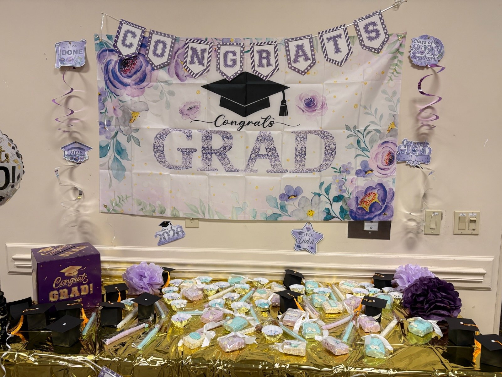 Decorative graduation celebration display with a banner saying 'Congrats Grad' featuring purple and white colors, floral accents, and graduation-themed decorations including caps, candies, and a box of graduation treats.