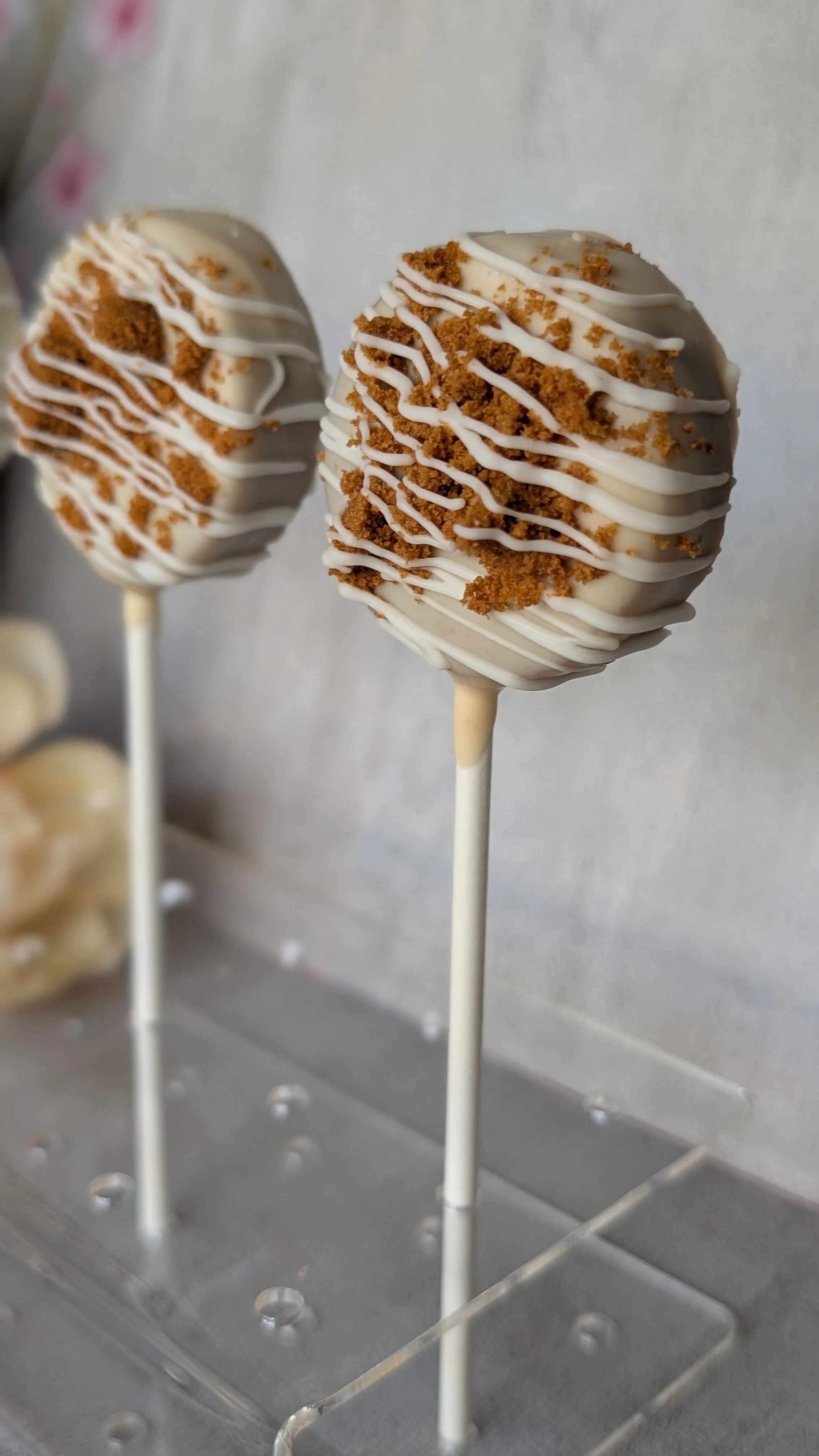 Two cake pops covered in white chocolate with crumbled cookie pieces and drizzled white chocolate on top, placed on silver stands.