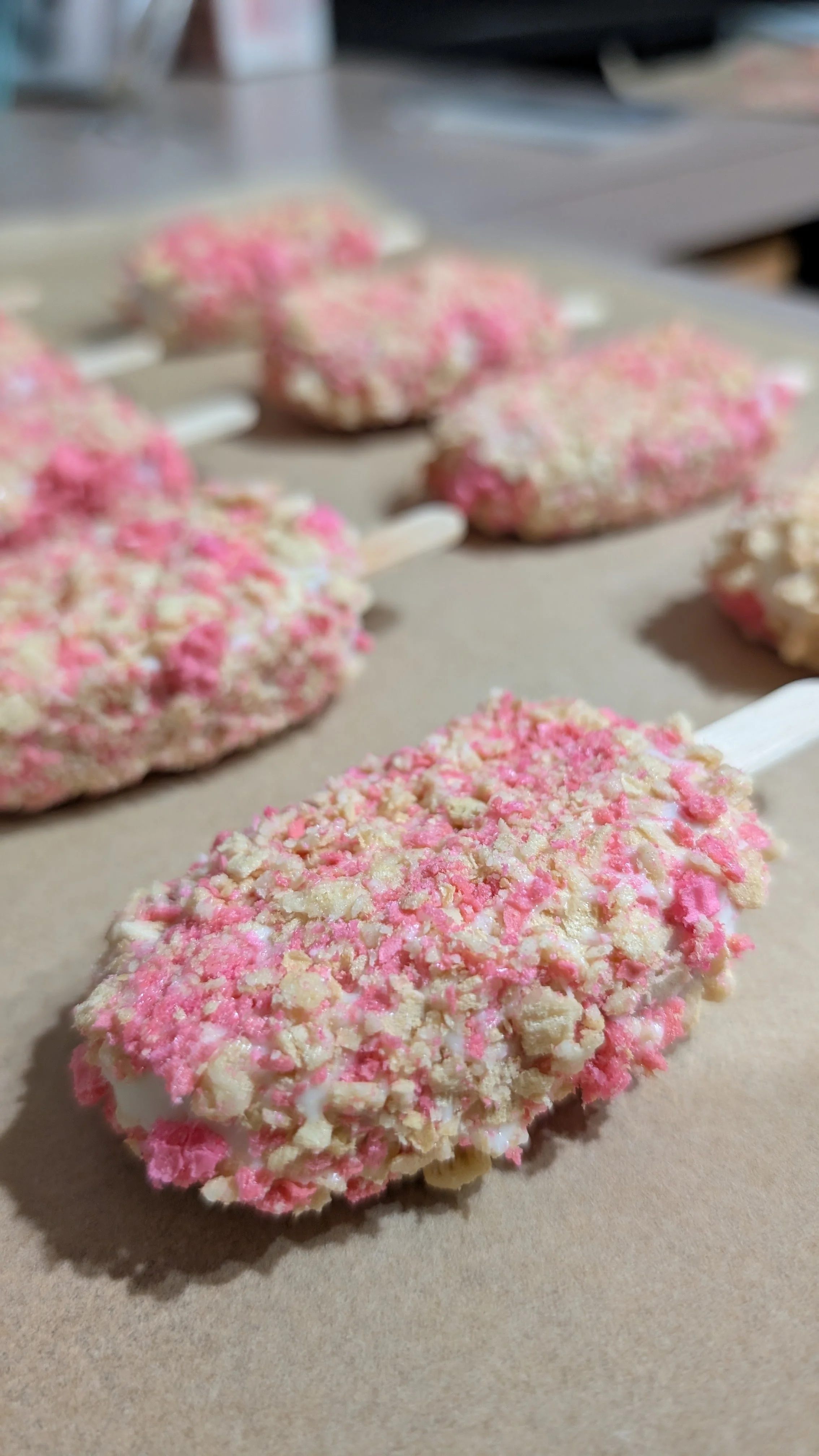 Close-up view of strawberry crunch-flavored cakesicles with pink and white sprinkles on a beige surface.