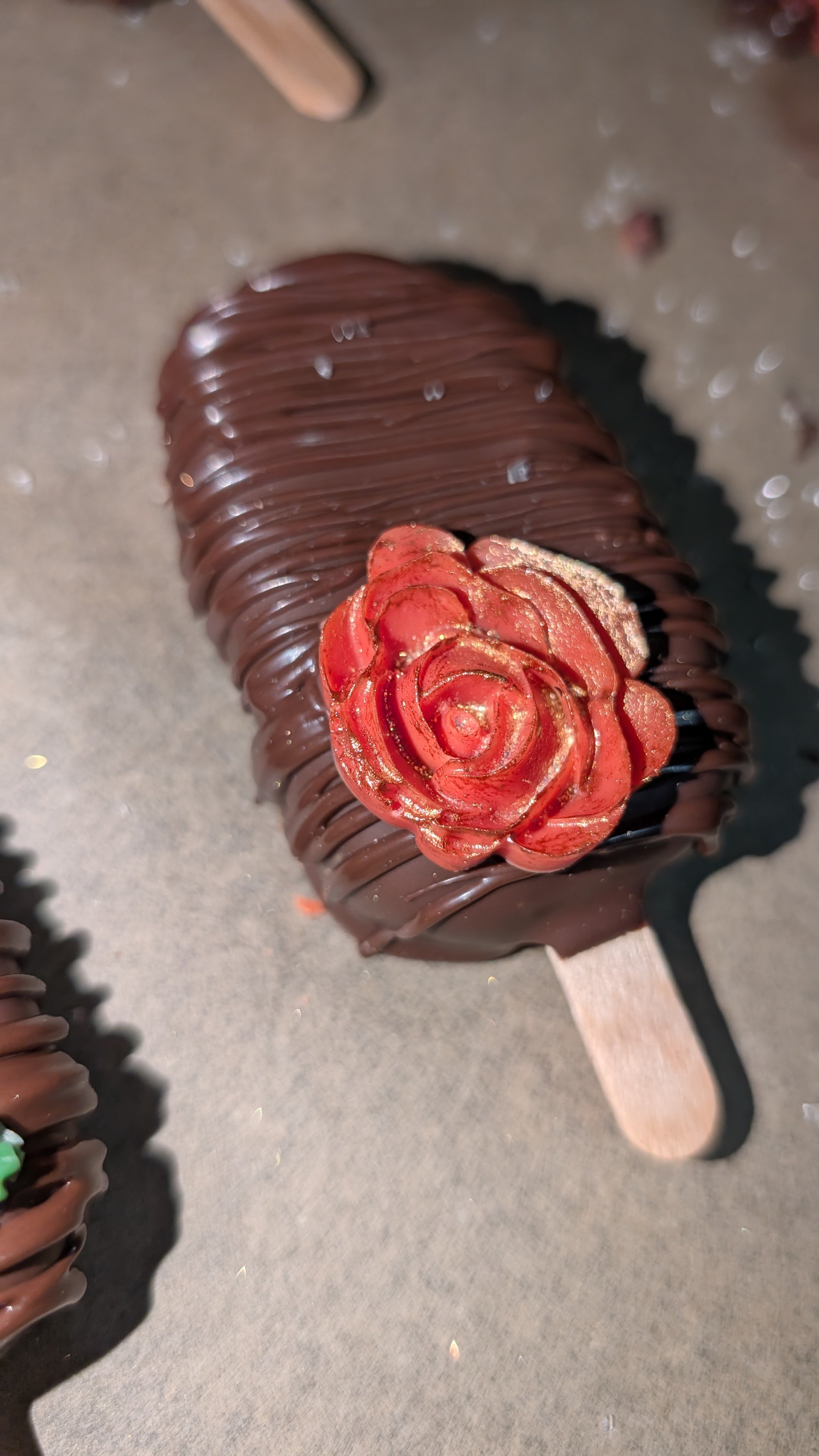 Chocolate covered strawberry cakesicle with a decorative red rose on top, on a wooden stick.