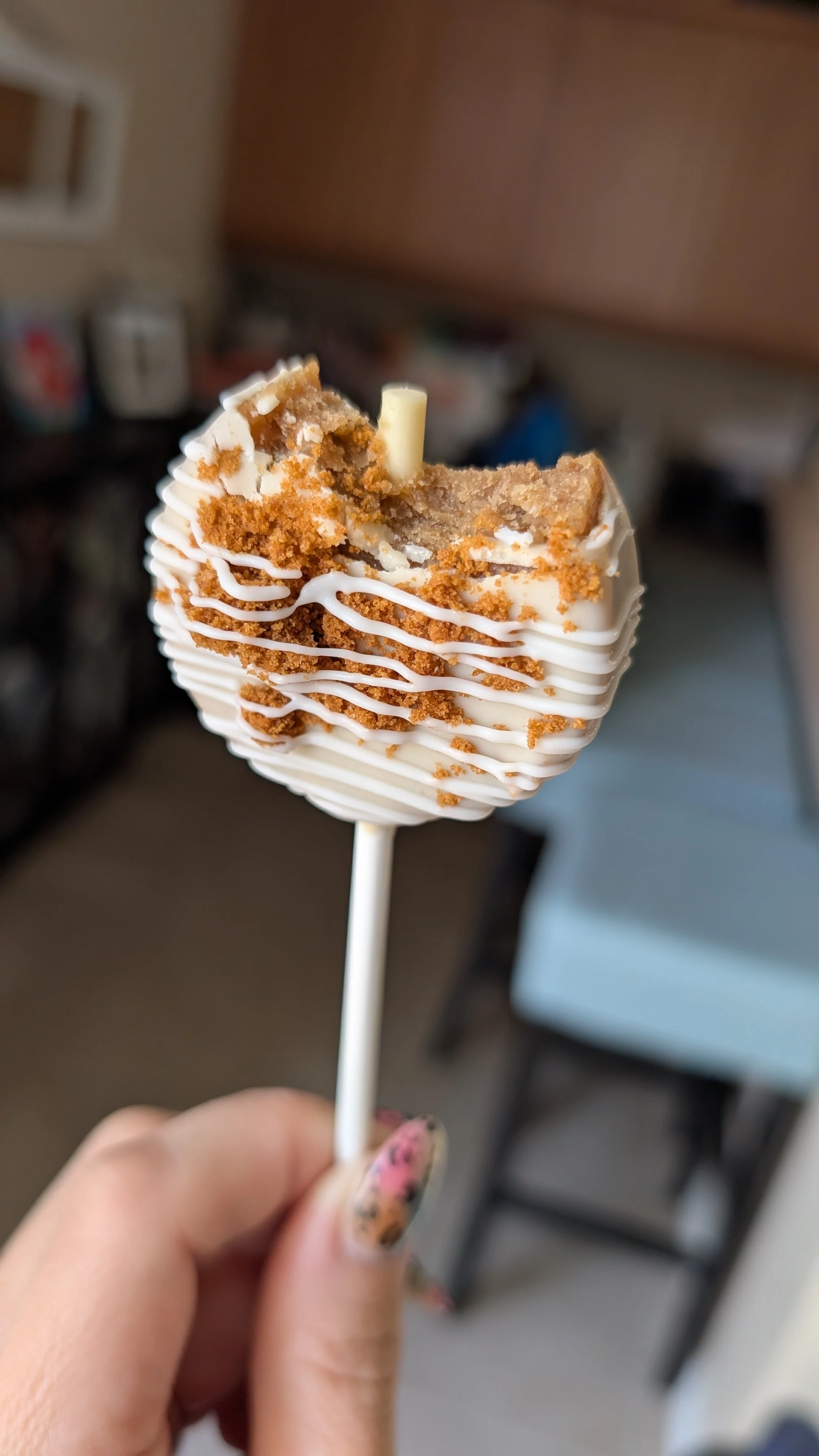 Close-up of a partially eaten biscoff cake pop with white icing, crumbled cookie or cake pieces, and white chocolate drizzle.