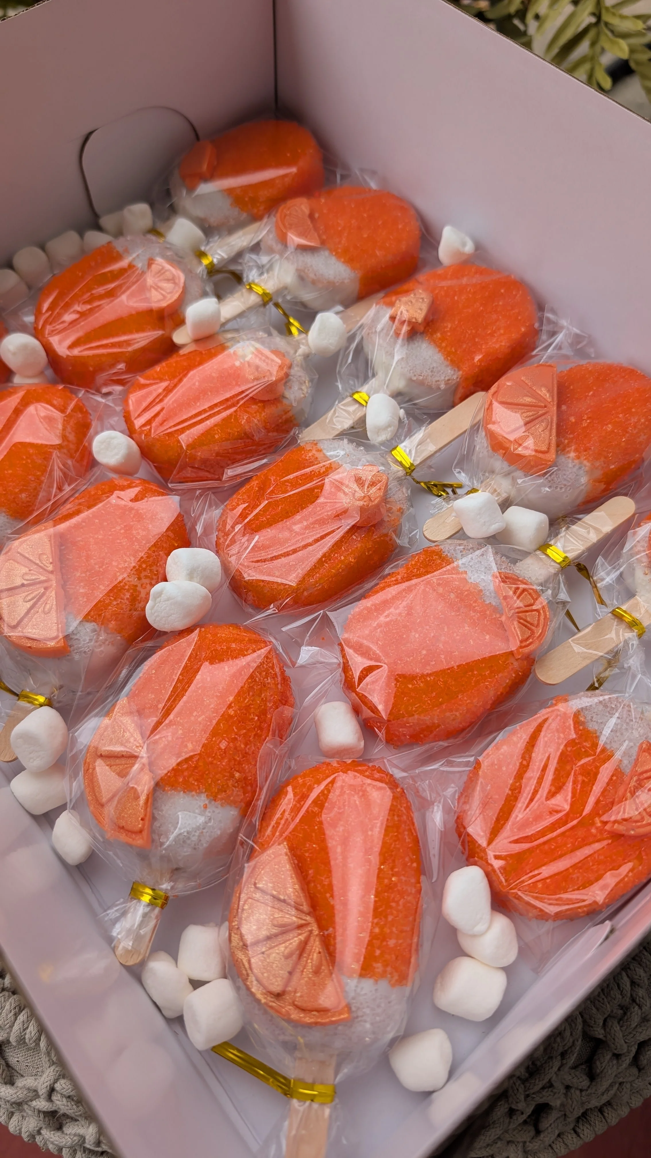Orange creamsicle cakesicles wrapped in clear plastic with small white marshmallows scattered around in a white box.