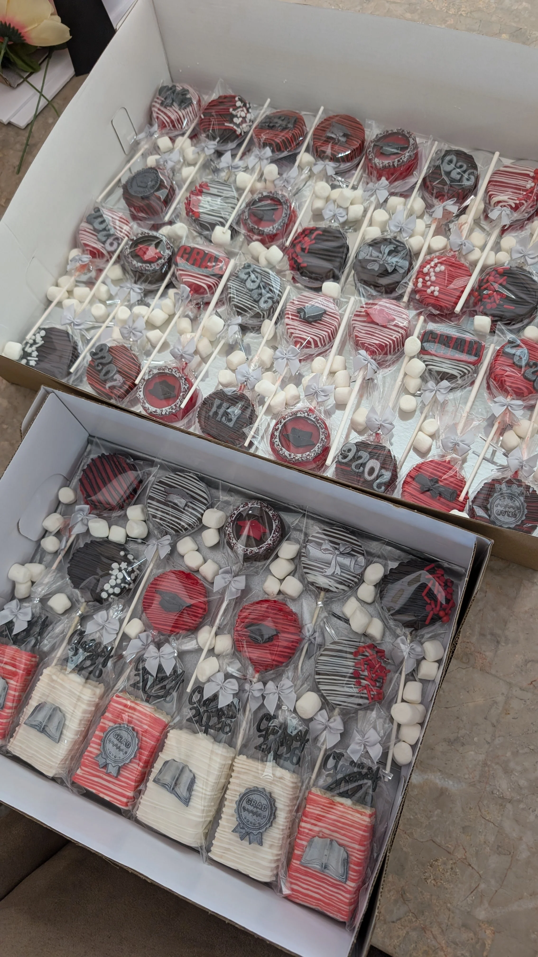 Box of decorated cookies and marshmallows for graduation celebration, with red, black, and white themes and graduation cap designs.
