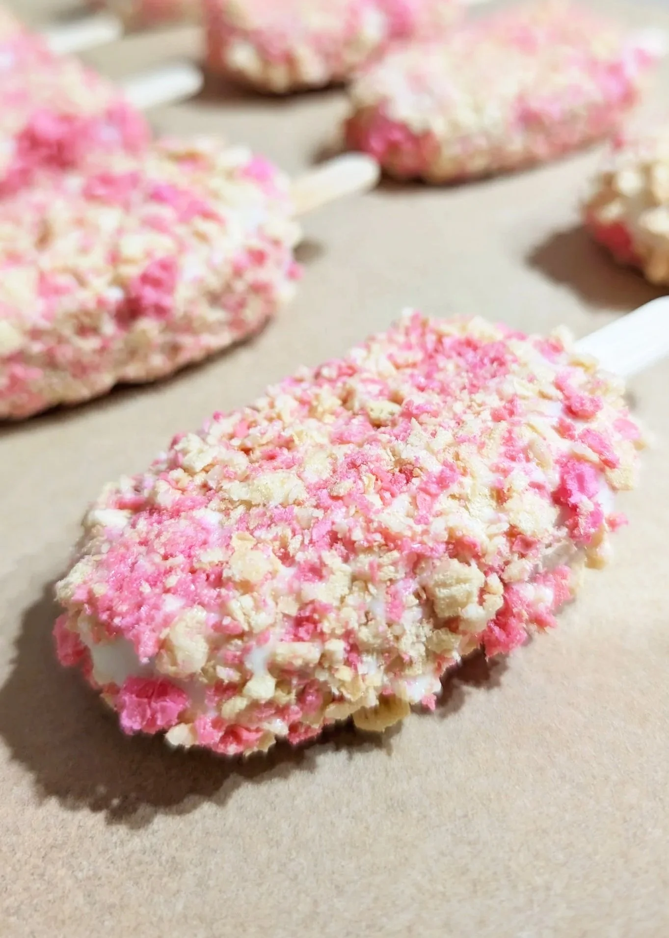 Close-up of pink and white ice cream bars covered with crushed pink and white toppings on a beige surface.