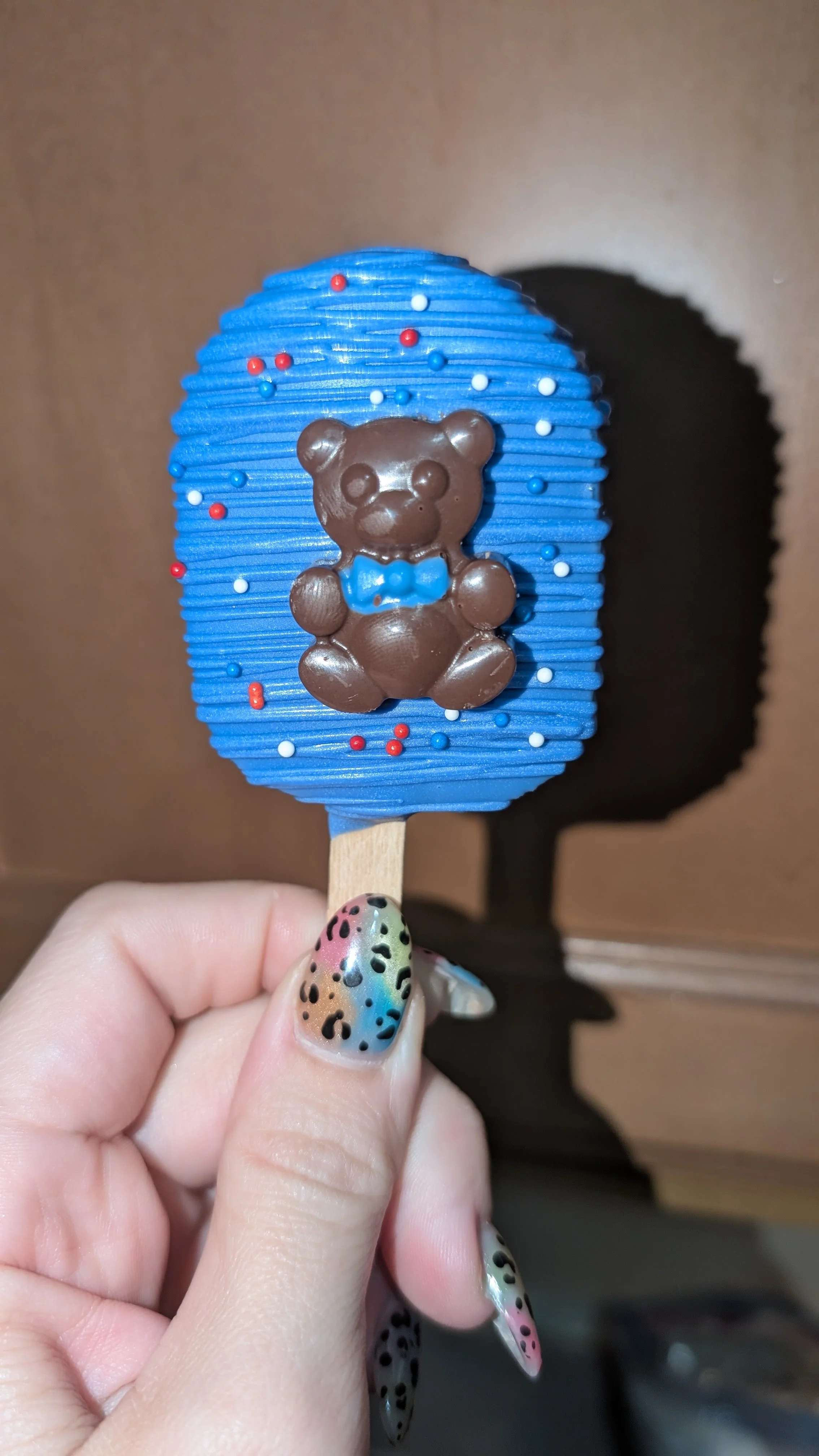 Blue popsicle decorated with multicolored sprinkles and a brown chocolate bear-shaped figure with a blue bow tie on top.