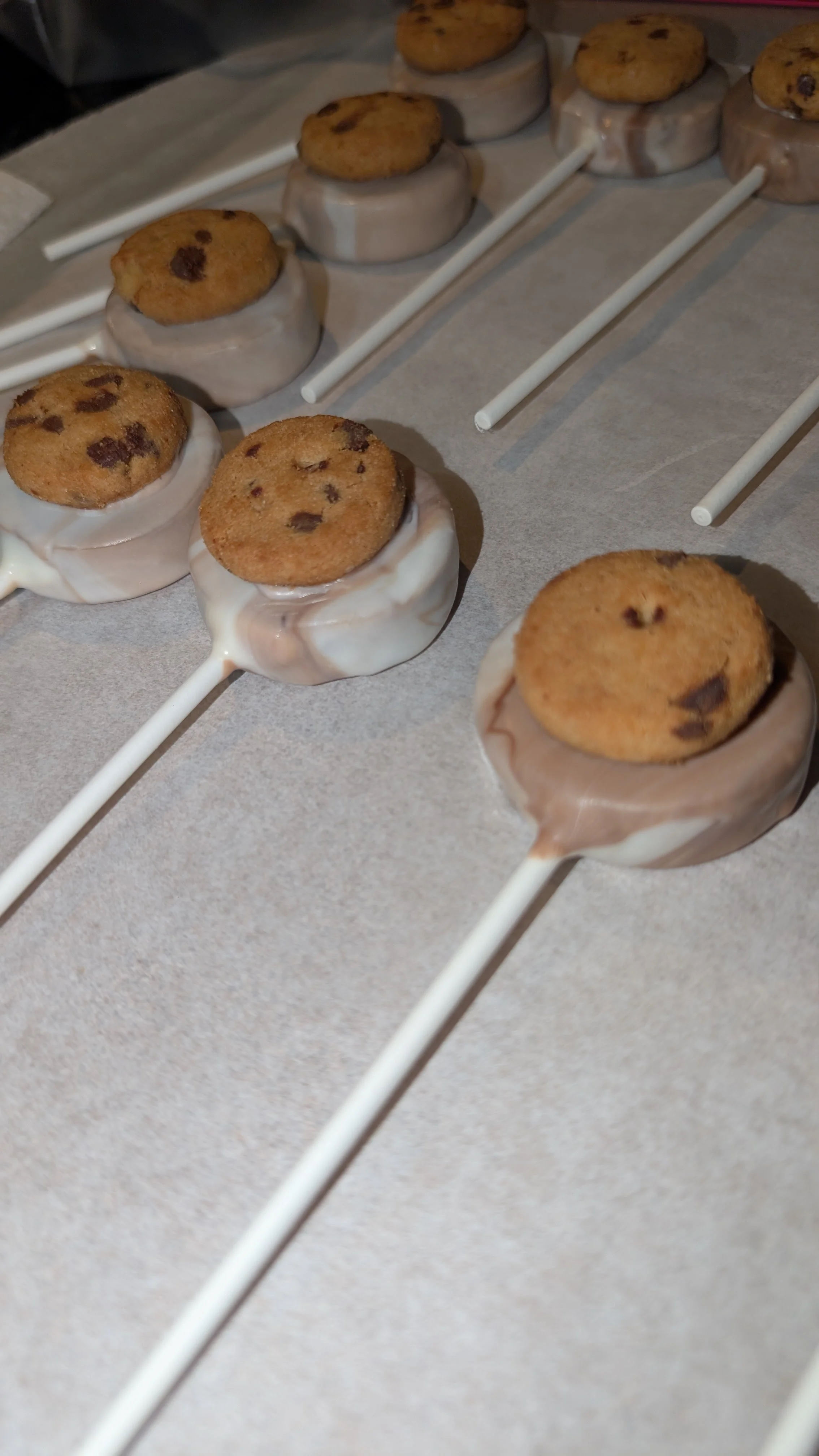 Chocolate chip cookie pops dipped in white chocolate on white sticks, arranged on parchment paper.