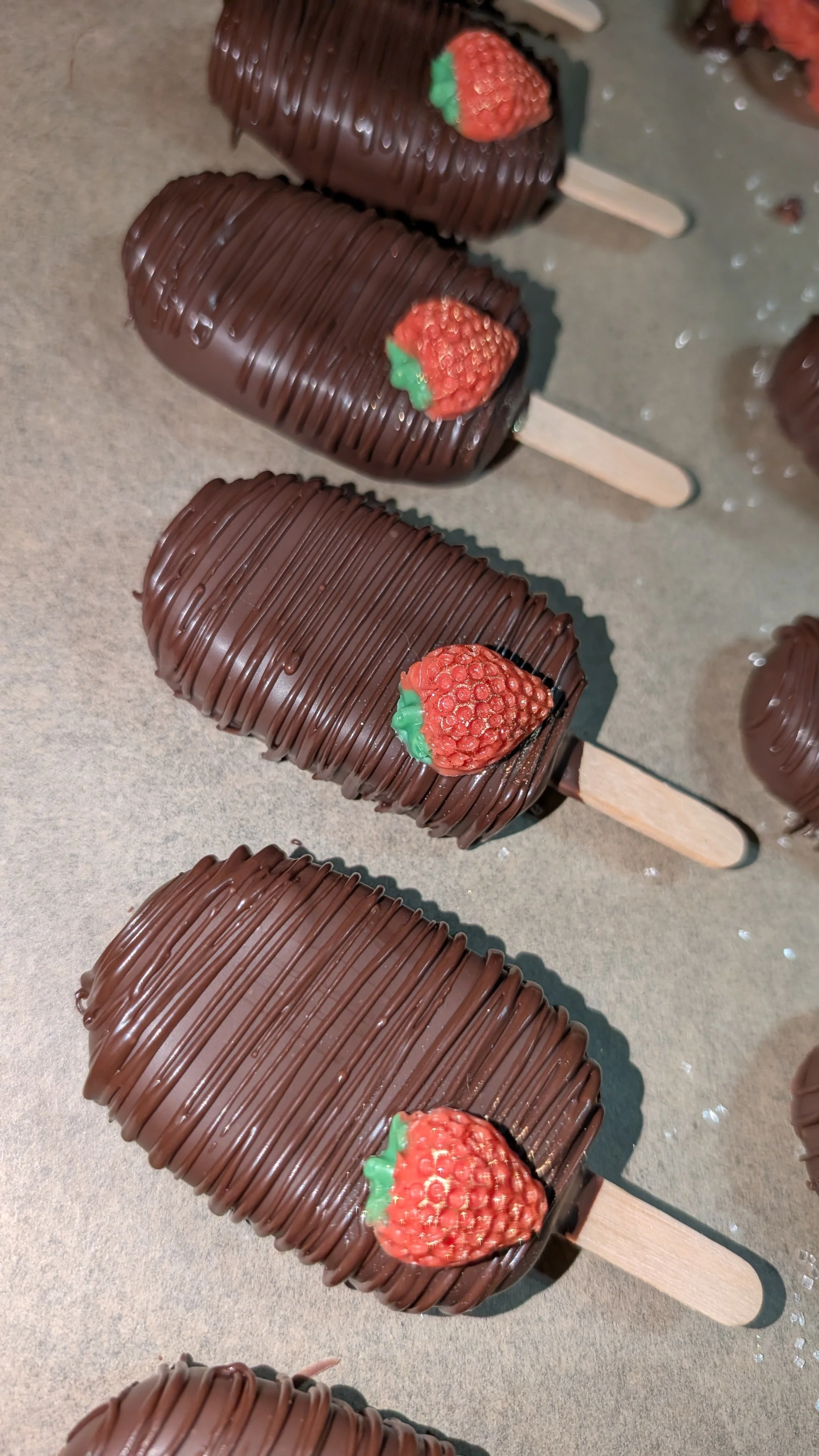 Chocolate-covered strawberry cakesicles with a strawberry and green decoration on top, lined up on a surface.