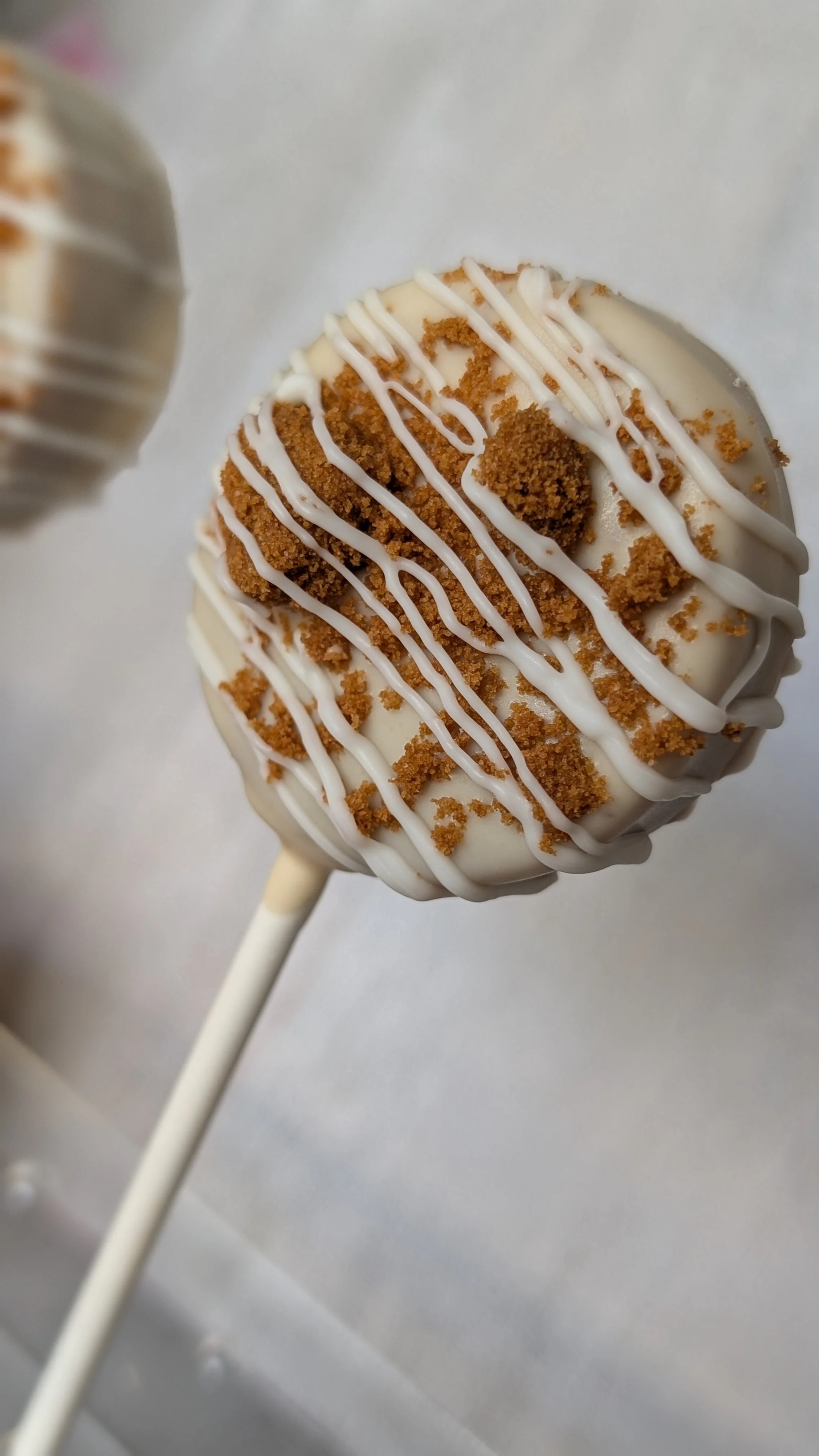 Close-up of a decorated biscoff cake pop with white icing, crumbled cookie topping, and white icing drizzle on a stick.