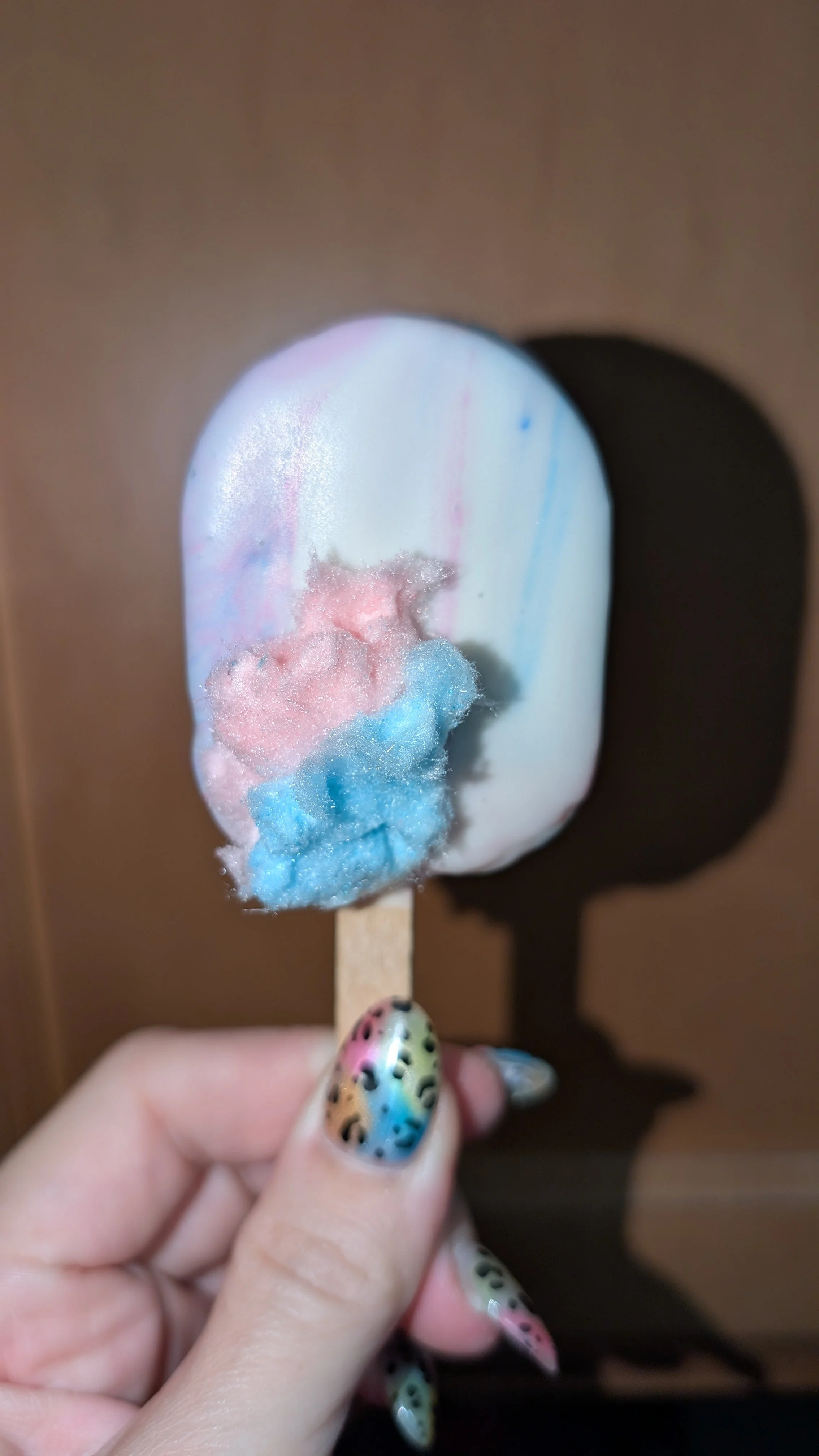 Close-up photo of a cotton candy flavored cake pop on a stick with pastel blue, pink, and white colors, held by a hand with colorful, patterned nails.
