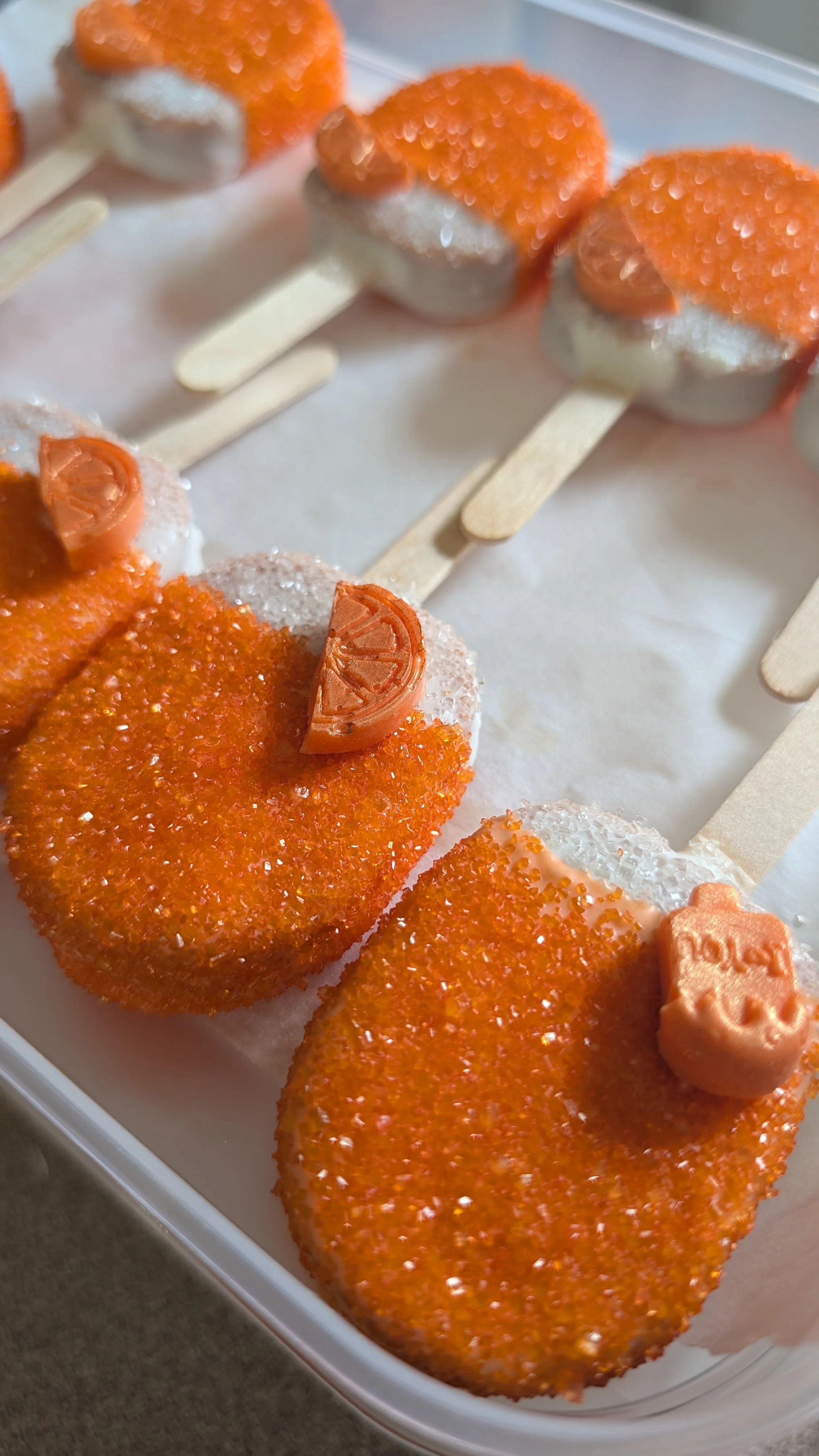 Close-up of orange creamsicle cake pops on wooden sticks with orange candies on top, arranged in a white container.