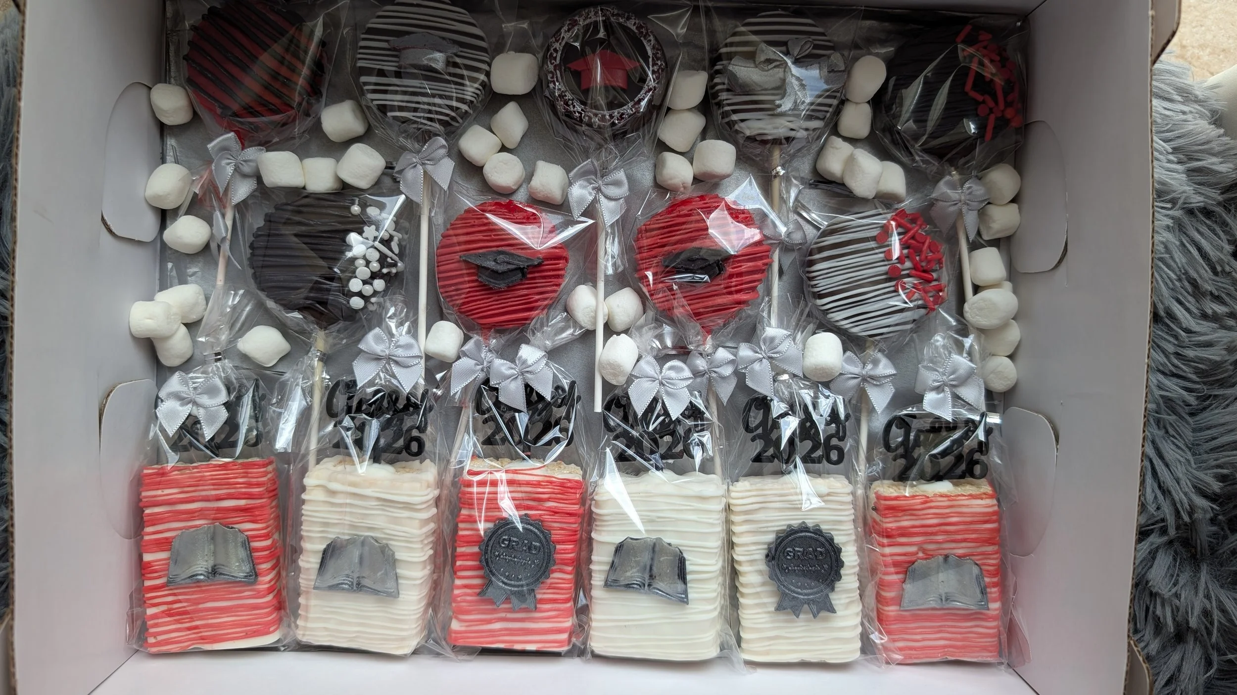 A collection of graduation-themed decorated cookies and marshmallows in a white box, with some cookies wrapped in clear plastic and tied with silver ribbons.