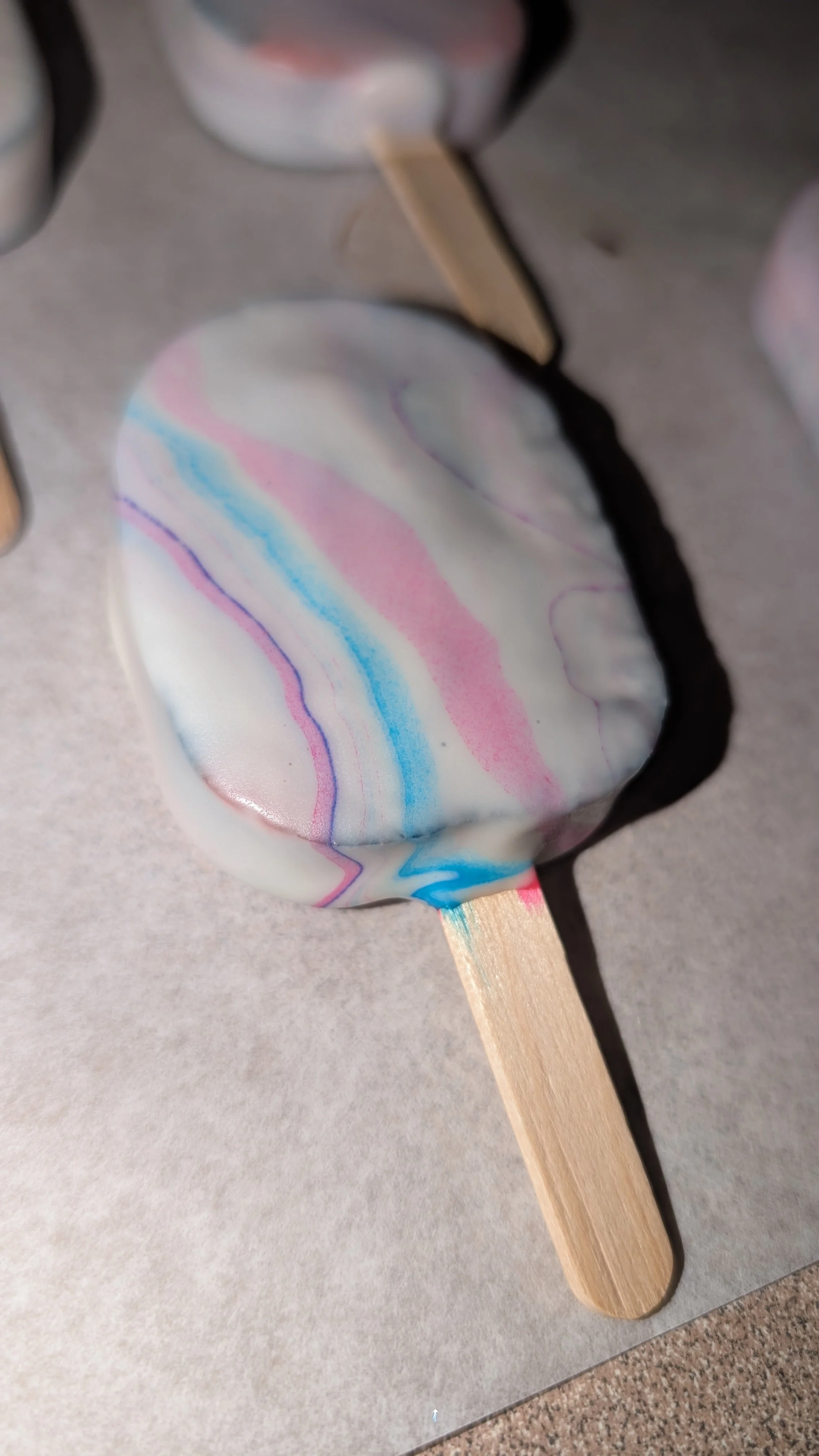 A heart-shaped cake with a marbled design of pink, blue, purple, and white icing on a wooden stick, placed on a gray surface.