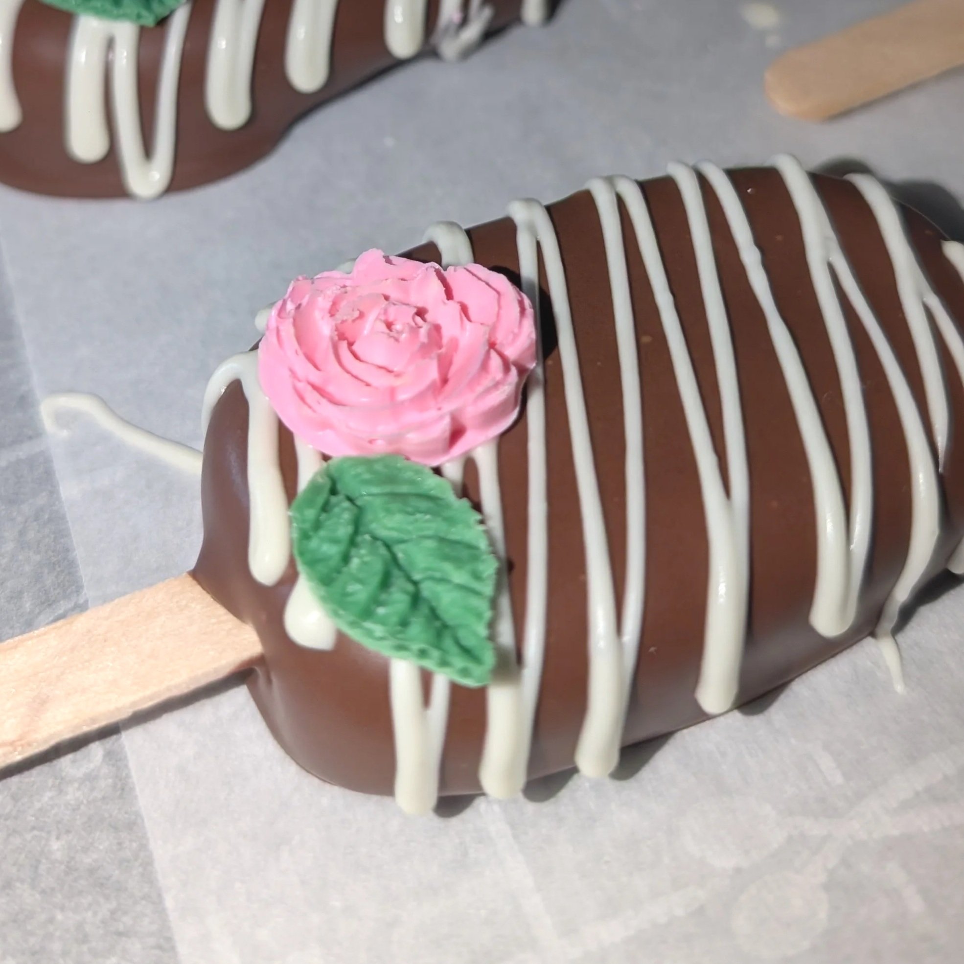 Close-up of a chocolate cake with white icing drizzle, topped with pink and green frosting decorations, on a plate.