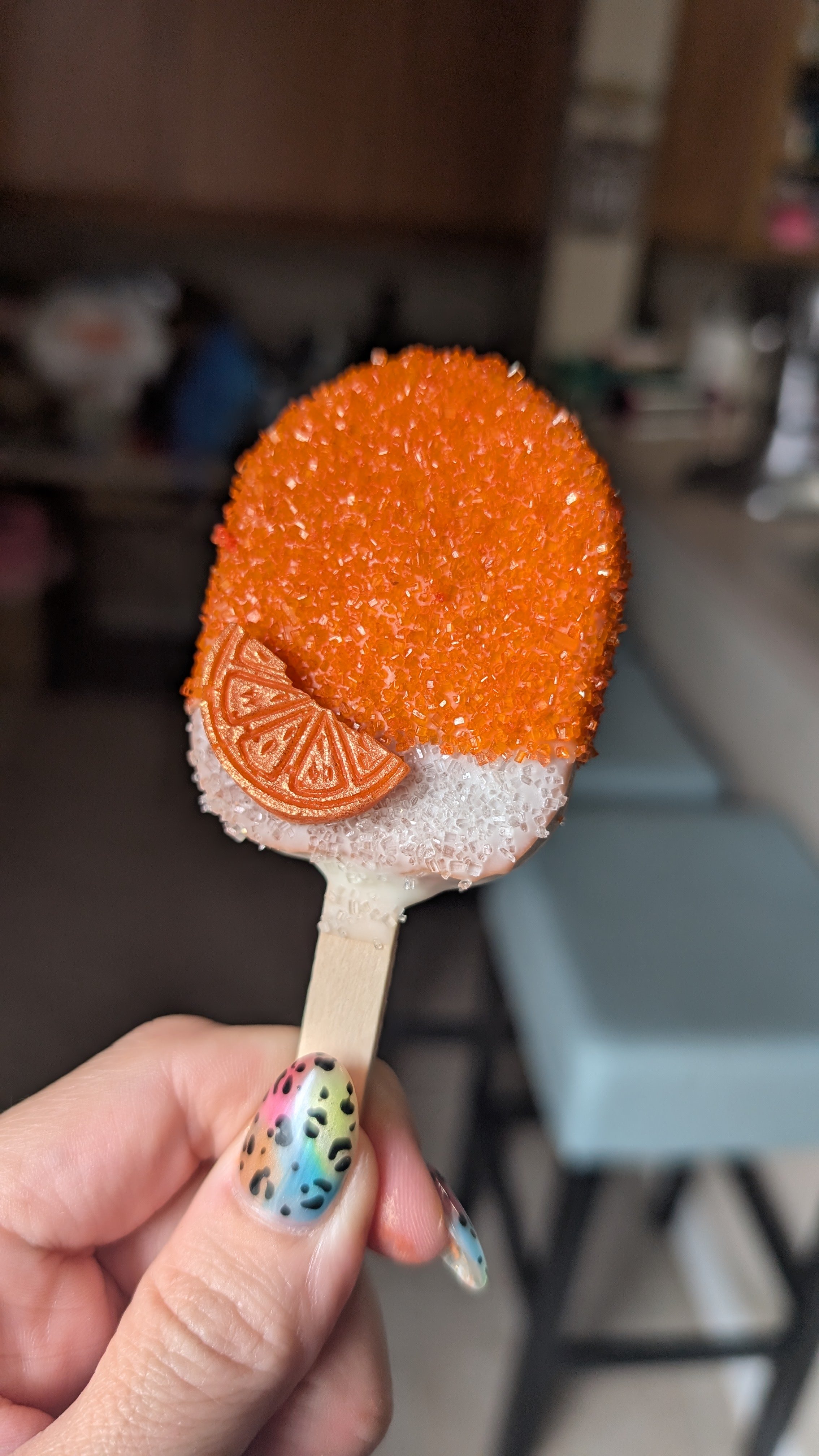 A hand holding a cakesicle decorated with orange and silver sugar crystals, a small orange slice, and rainbow-colored nail polish.