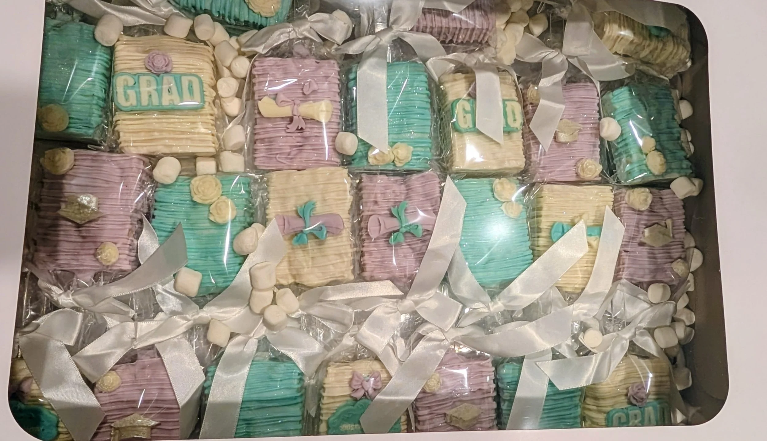 A box of decorated cookies in pastel colors with ribbons. Some cookies spell out 'GRAD', some have bows, and others have decorative flowers.