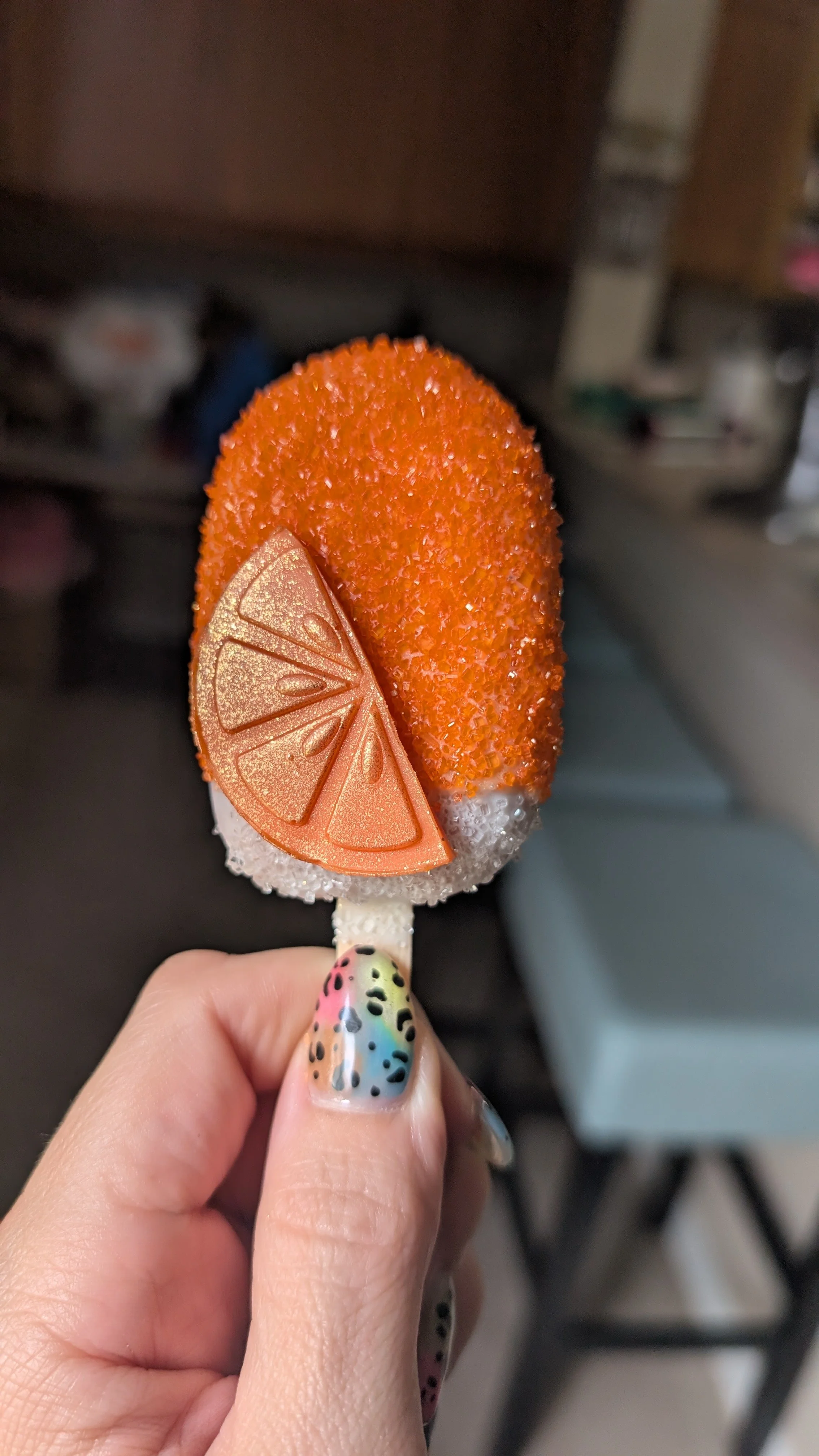 A person holding a cakesicle topped with orange and lemon slices decorations, with colorful nail art.