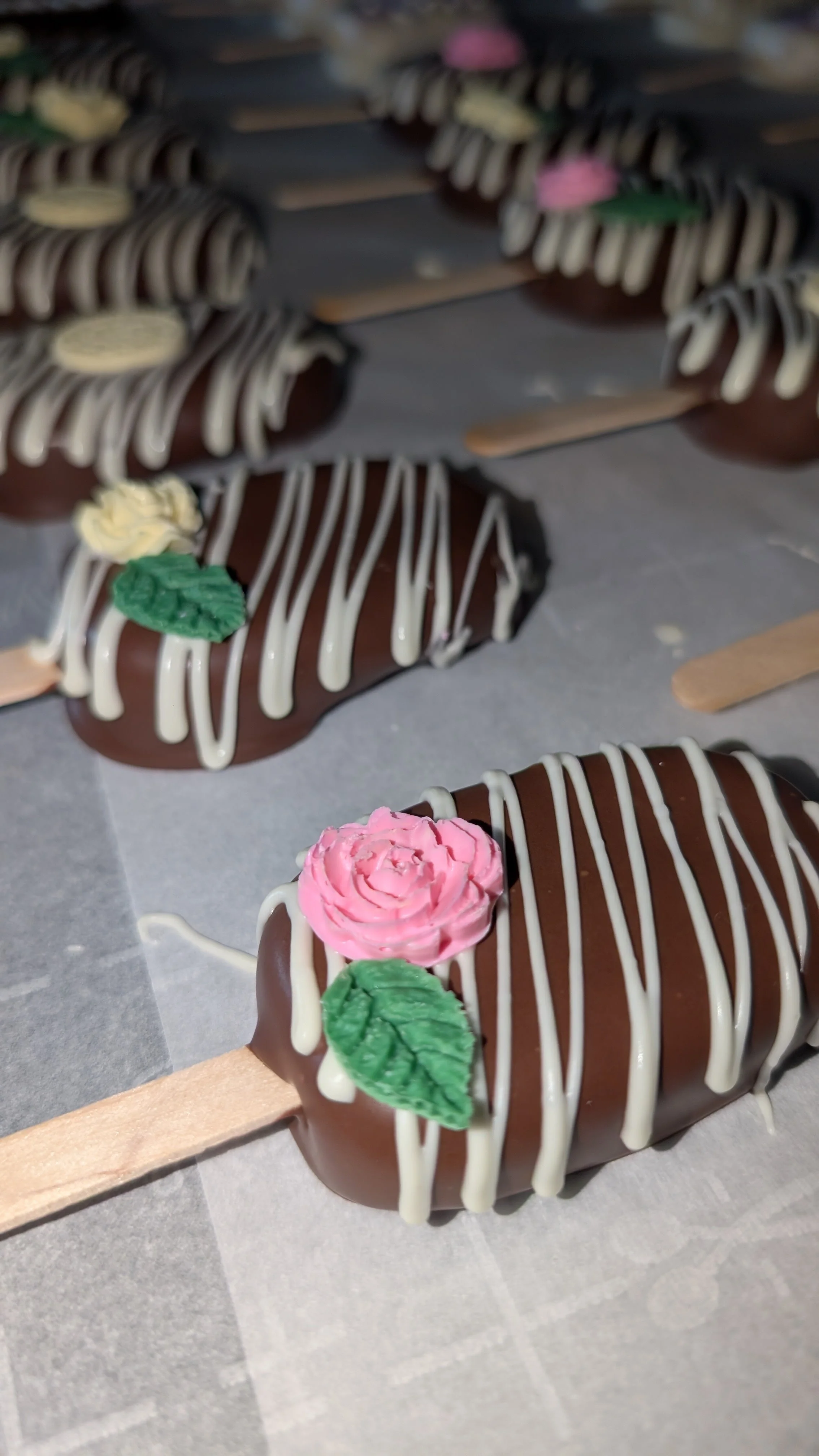Chocolate-covered ice cakesicles on sticks decorated with pink and green frosting flowers and white drizzle.