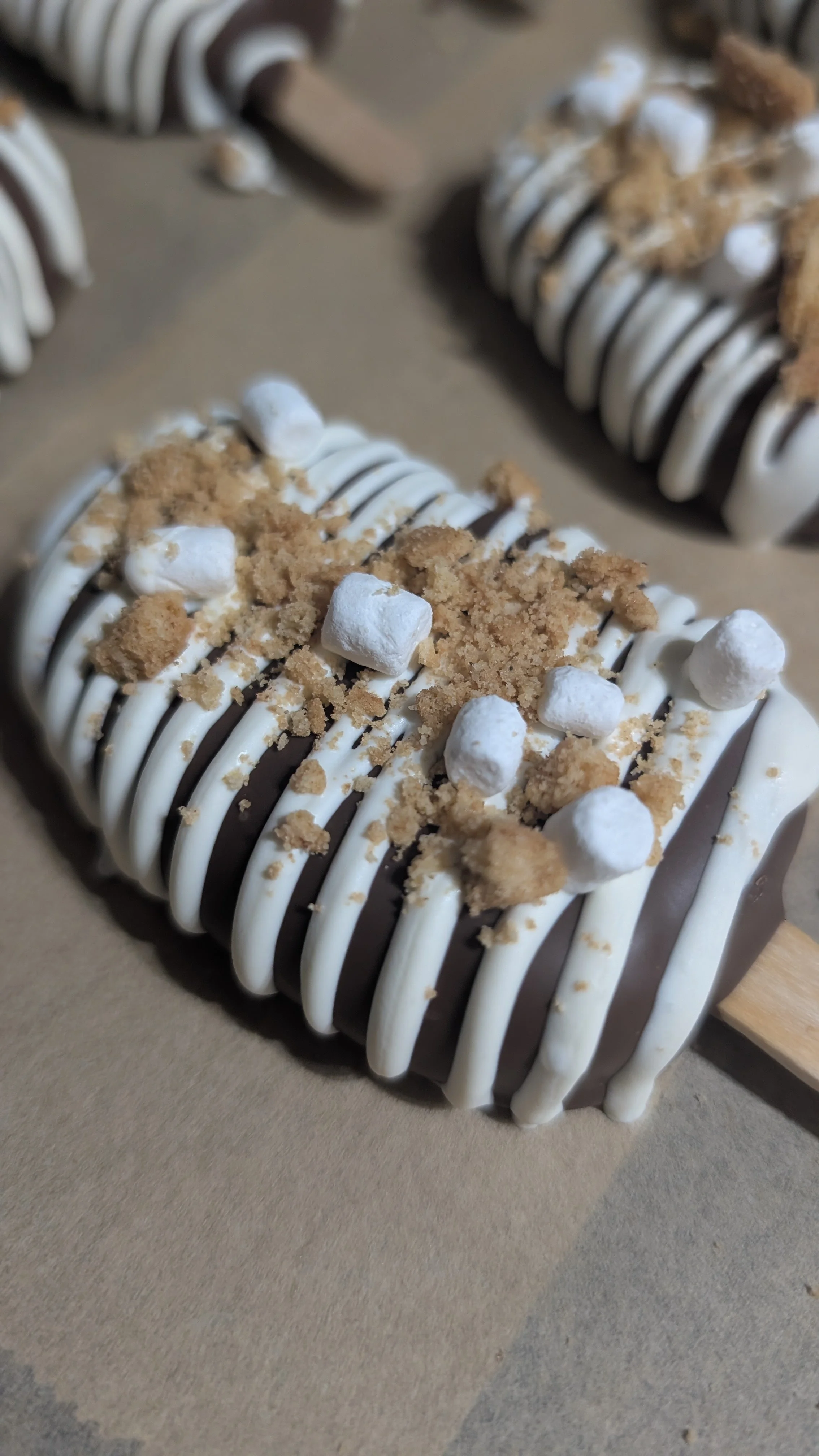 smores cakesicle with white chocolate drizzle, crushed cookies, and mini marshmallows.