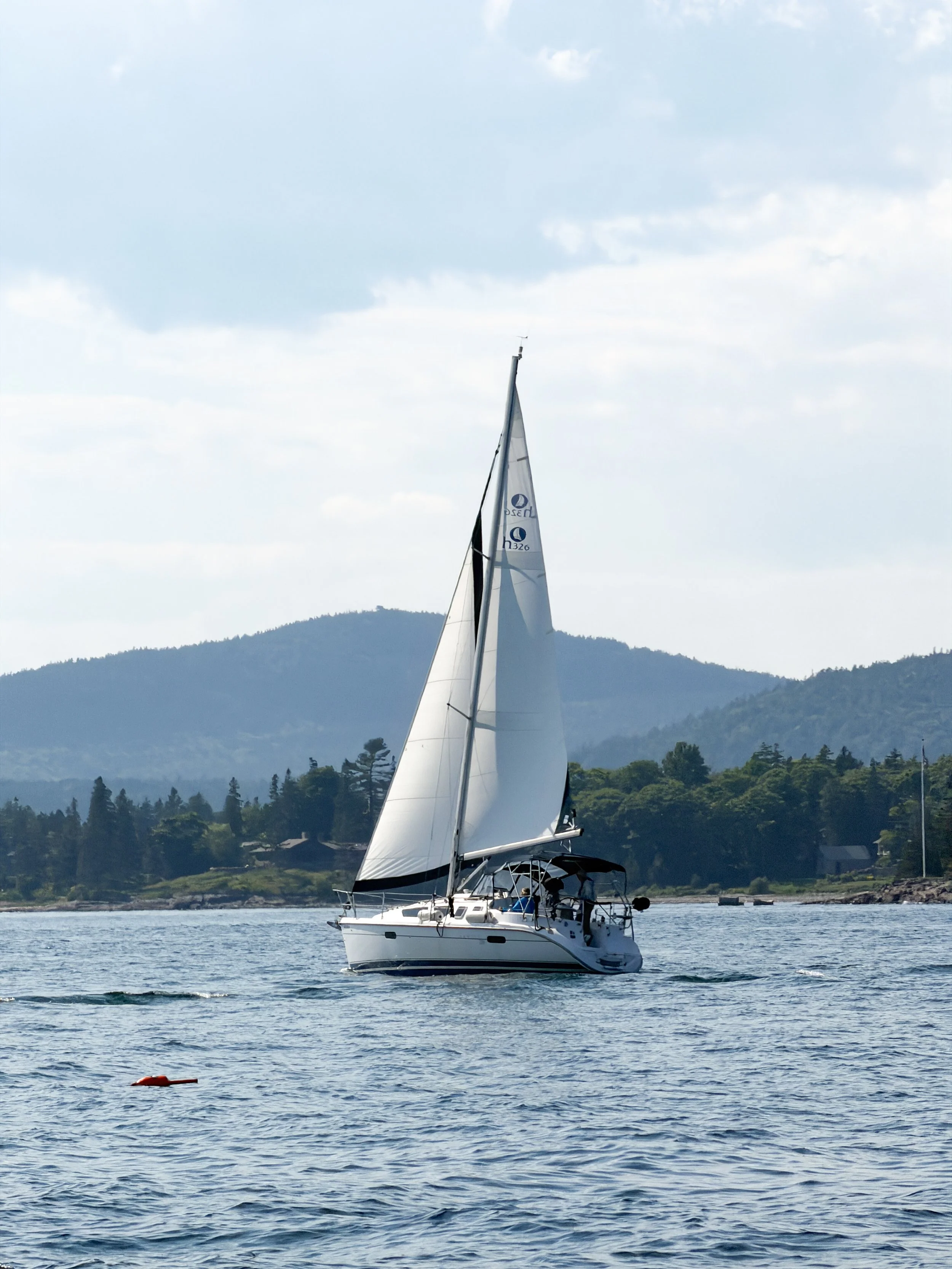 A sailboat with white sails on a body of water, with a mountainous landscape and partly cloudy sky in the background.