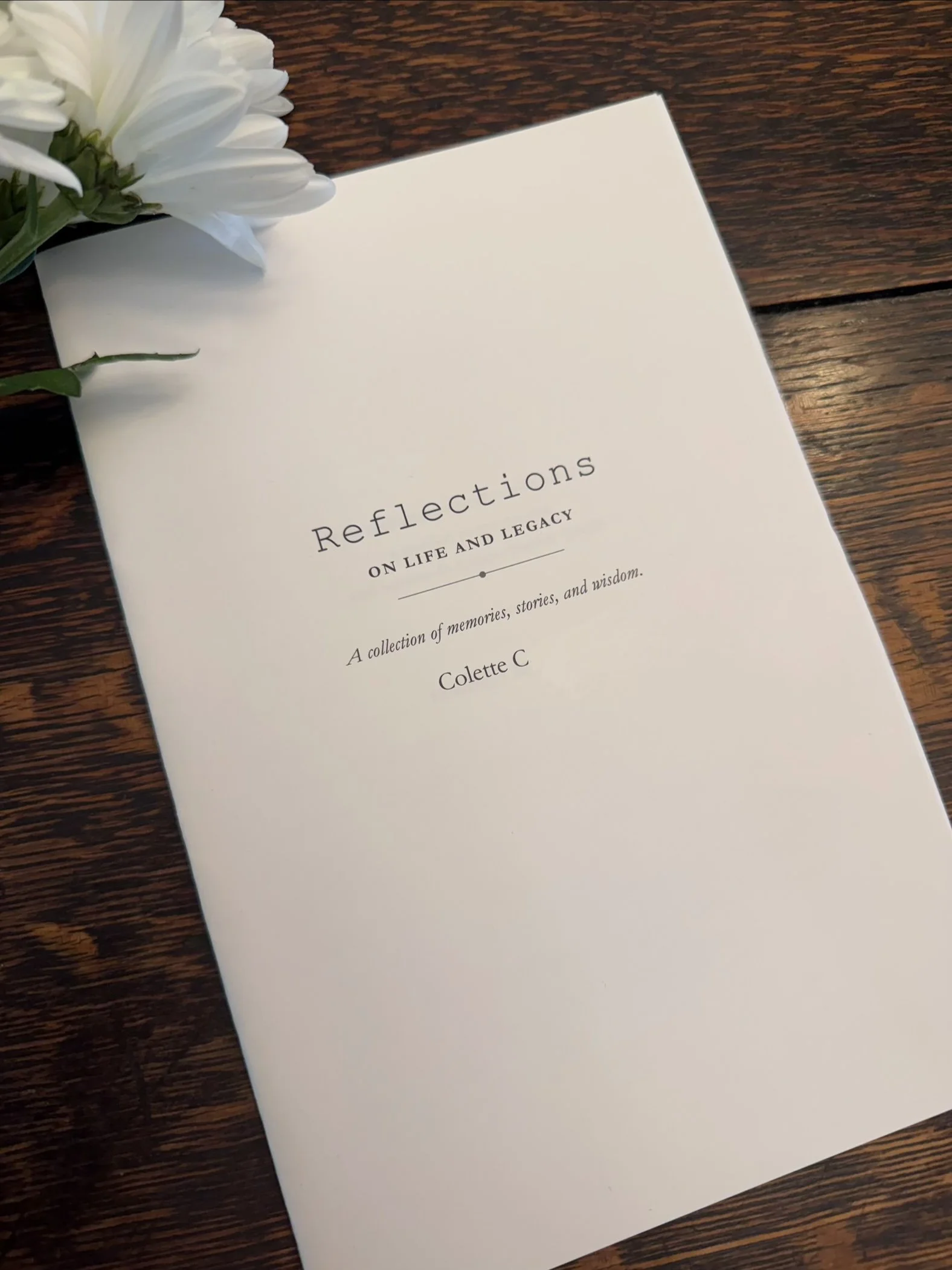 A white book titled 'Reflections on Life and Legacy' is placed on a wooden surface, with white flowers resting on top of it.