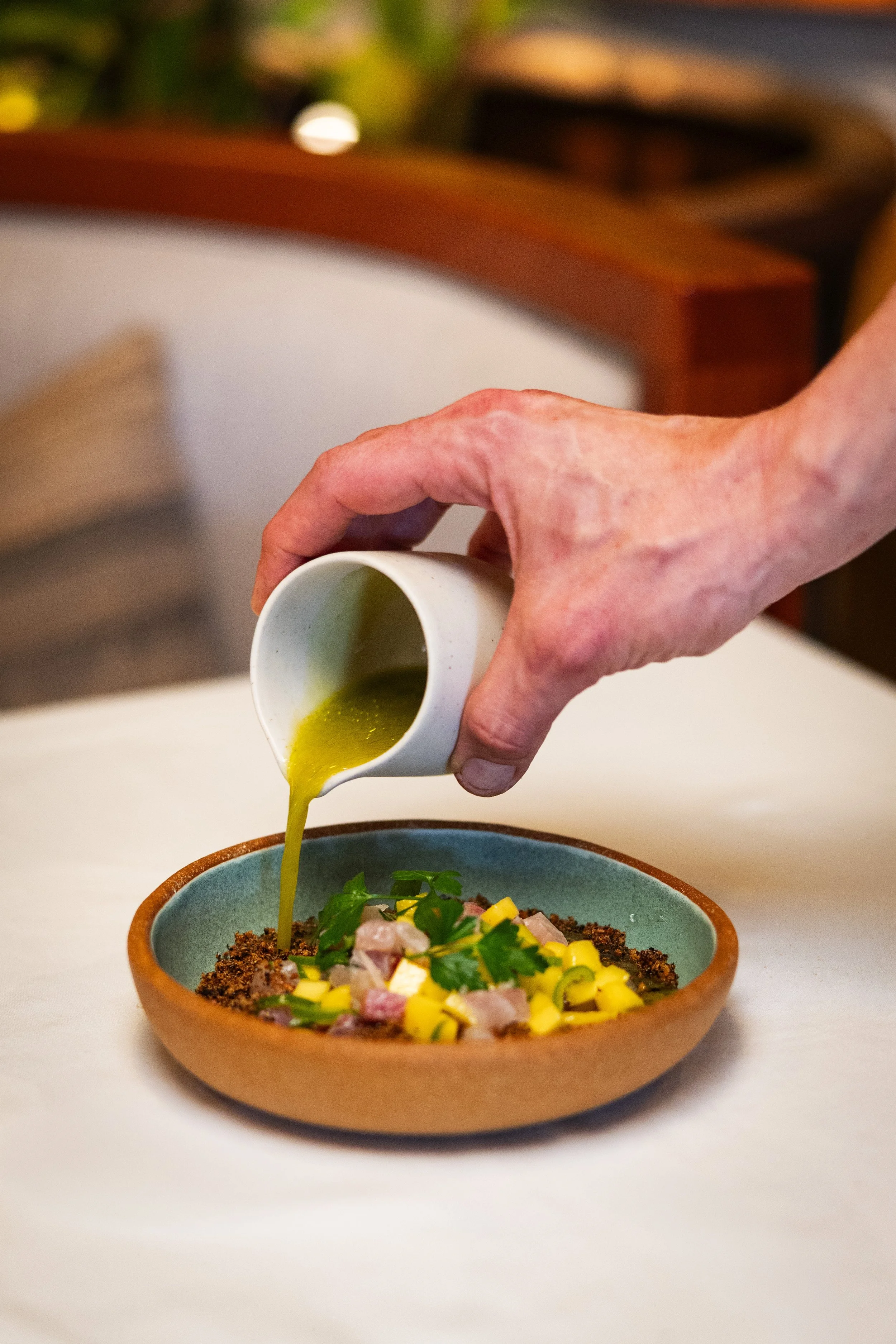 A person is pouring green sauce onto a colorful dish with chopped vegetables and herbs in a blue and brown bowl.