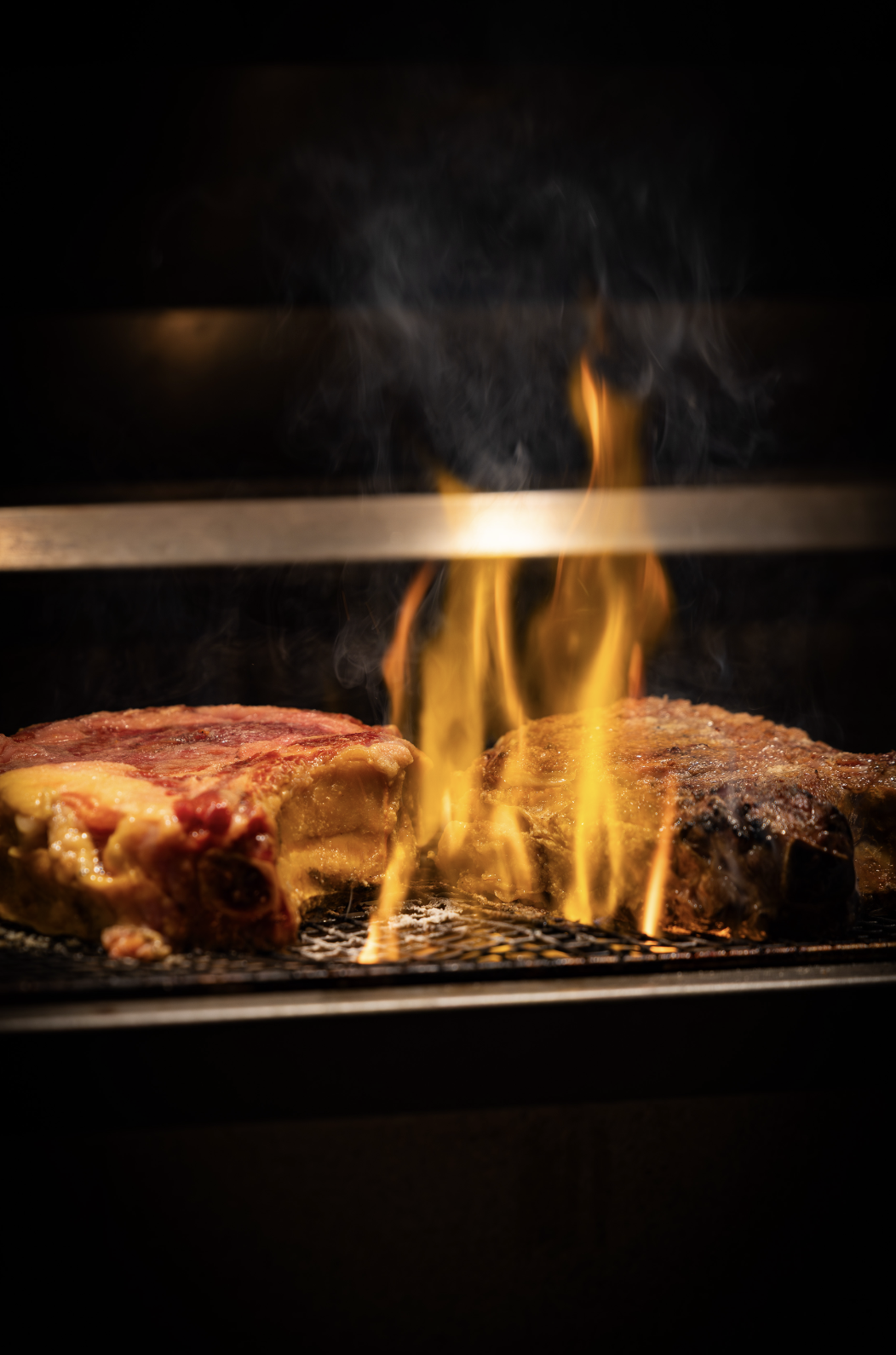 Close-up of two steaks cooking on a grill with flames and smoke rising, dark background.